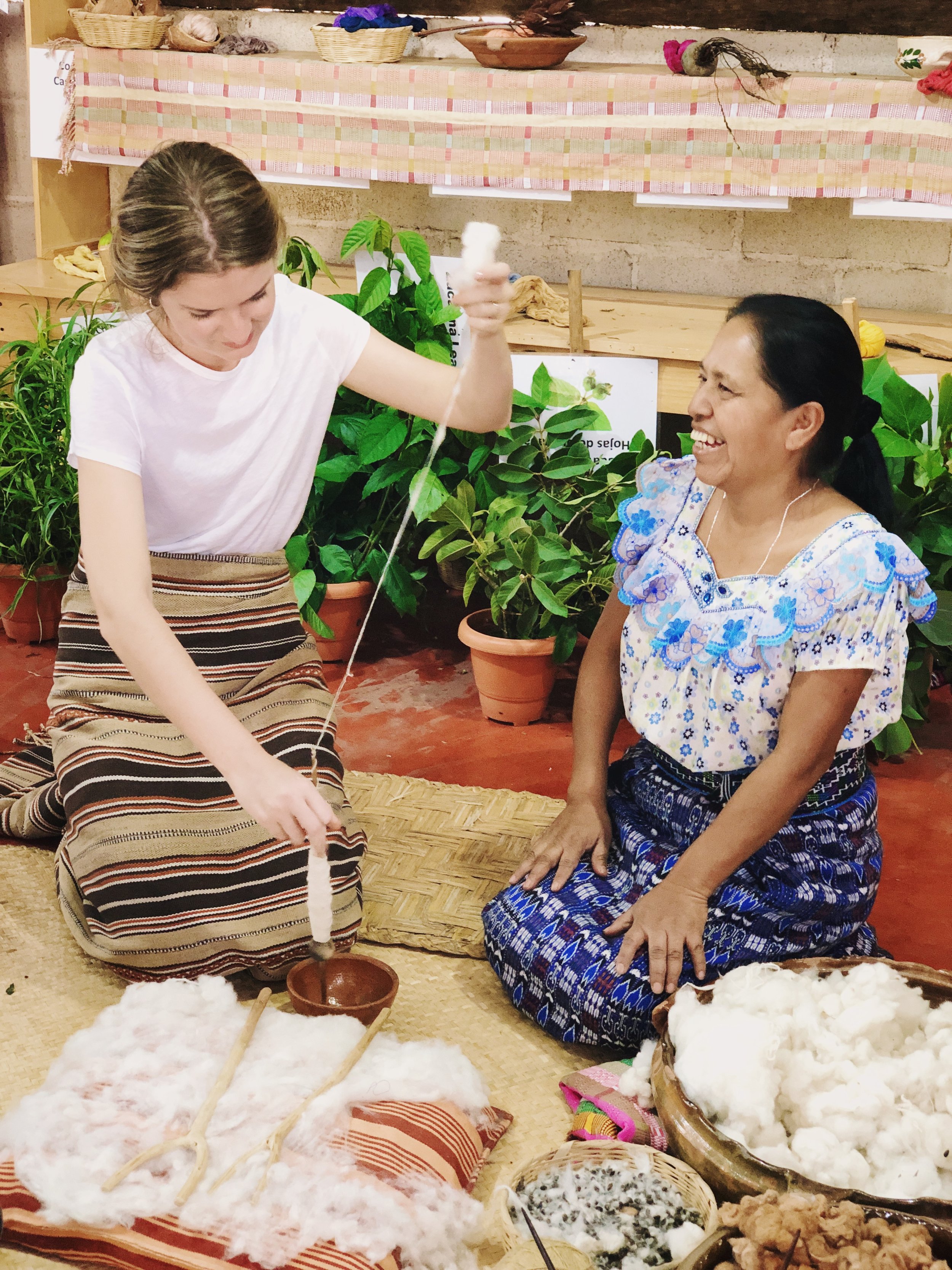 Young woman and older woman sitting on the floor surrounded by cotton plants and fibers, engaging in cotton processing, with green plants in the background.