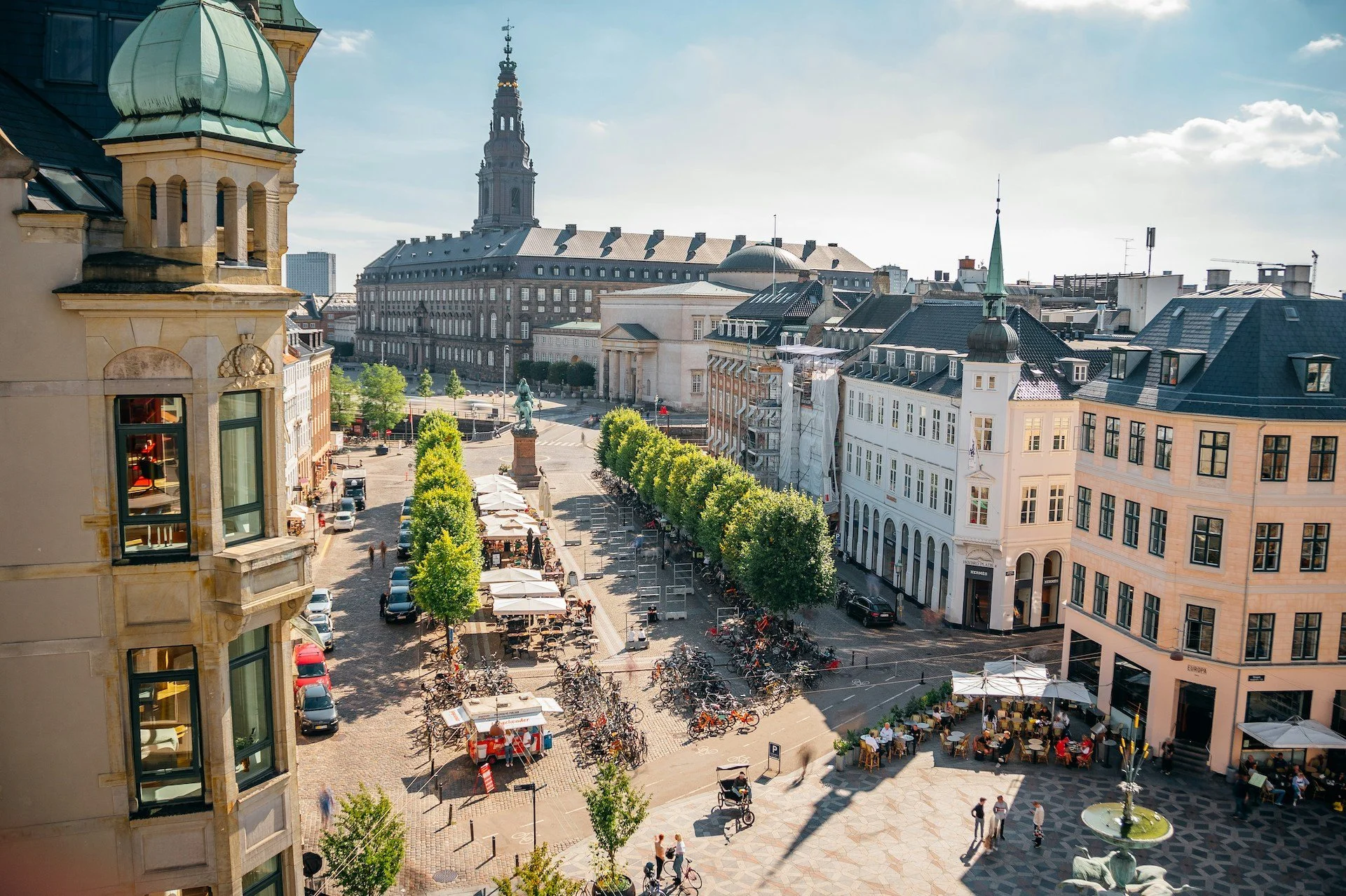 City square with outdoor cafes, bicycles, and pedestrians in front of historic buildings and a monument, with a large government building and spires in the background.