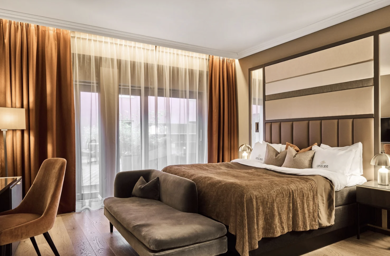 Luxury hotel room with large bed, beige upholstered headboard, bedside lamps, brown curtains, sheer curtains, brown bedding, decorative pillows, and seating area with a gray sofa and light brown chairs.