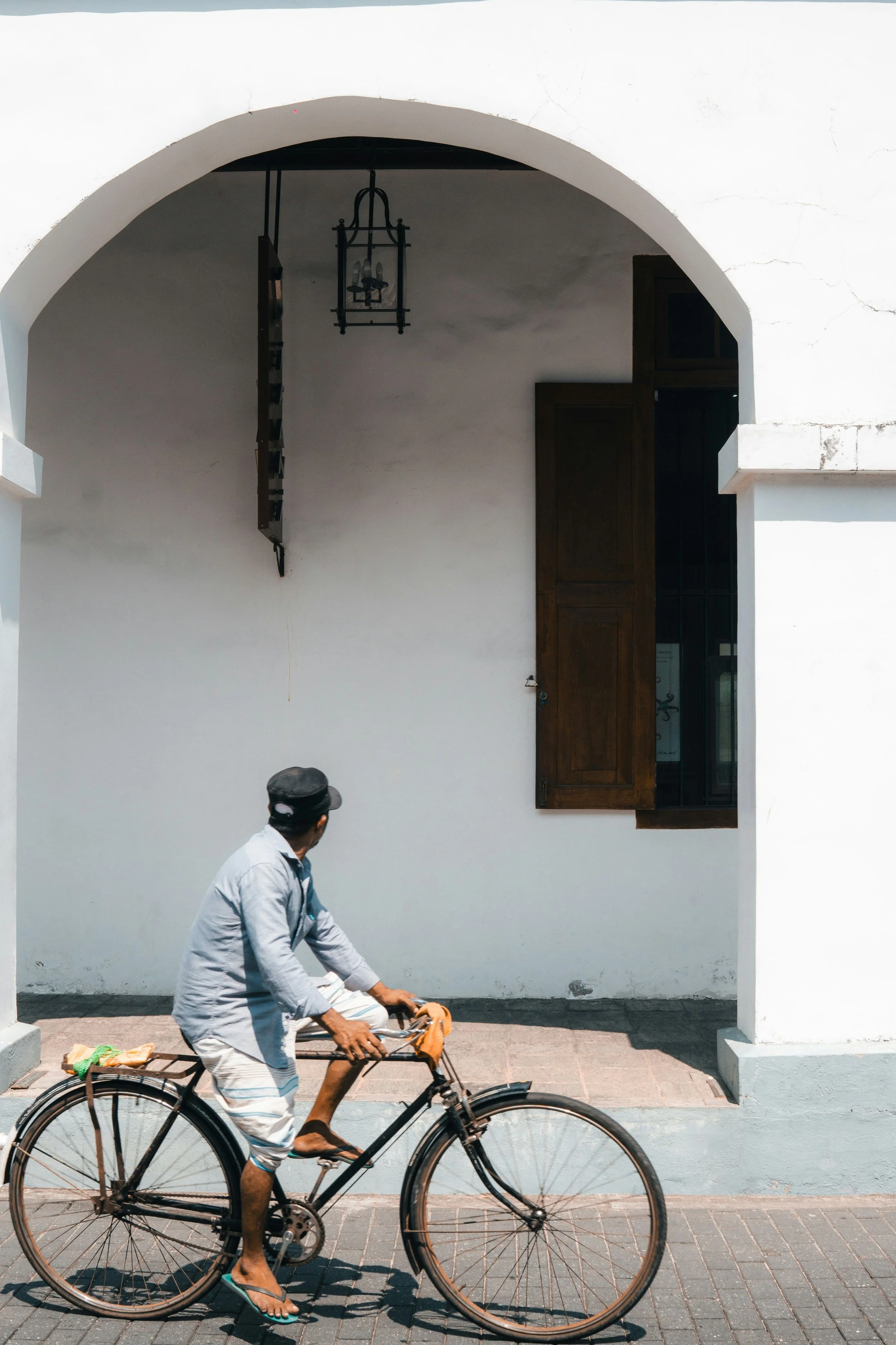 Man on vintage bicycle under white archway with wooden window and hanging lantern.