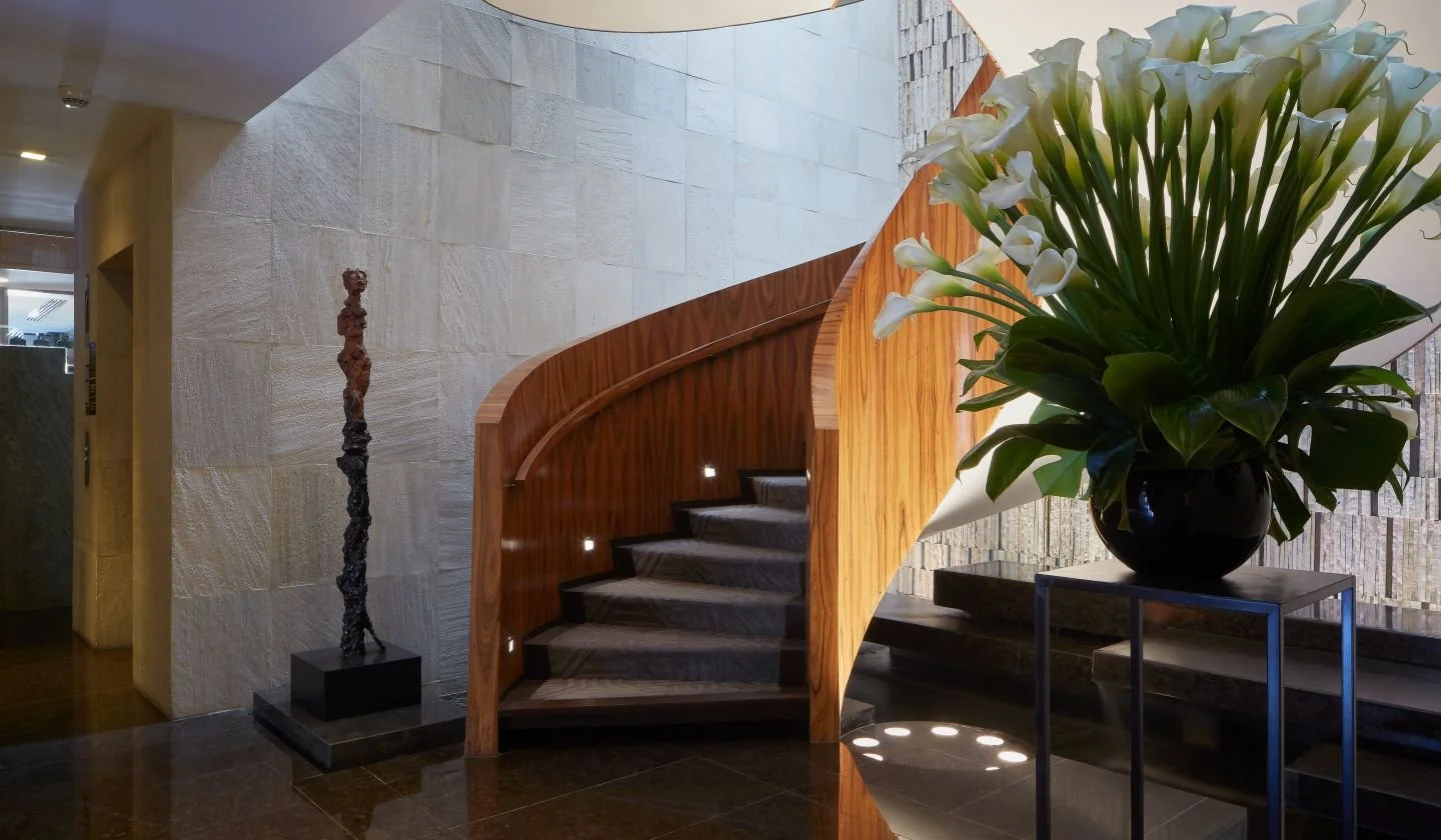 Elegant interior with a curved wooden staircase, large potted calla lilies, and a modern sculpture in a well-lit room.