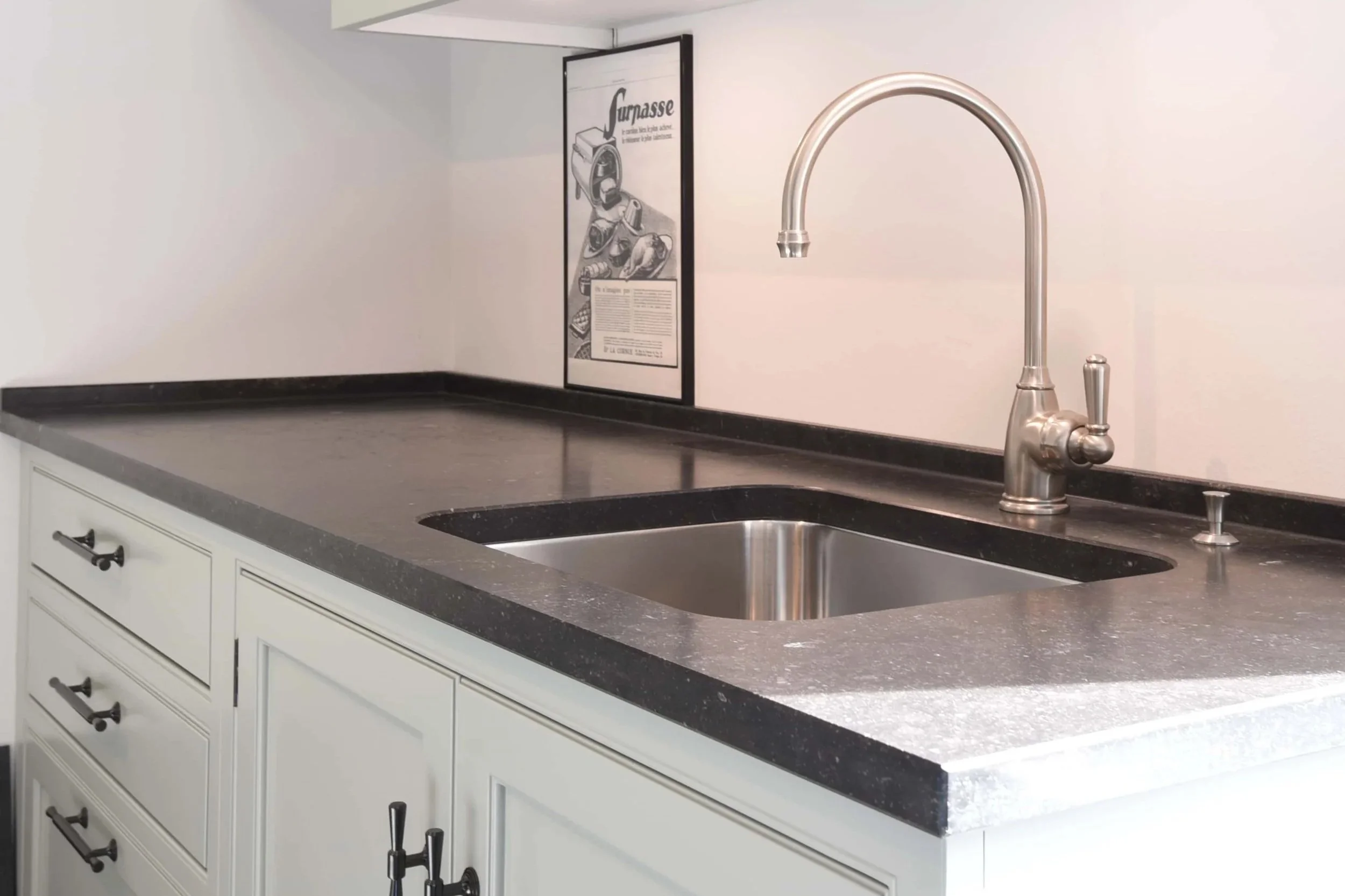 Küchenarbeitsplatte aus Blausstein, mit grünen Schränken darunter, Edelstahlspüle mit Wasserhahn in Edelstahl, schwarze Griffe, Beleuchtung von oben.