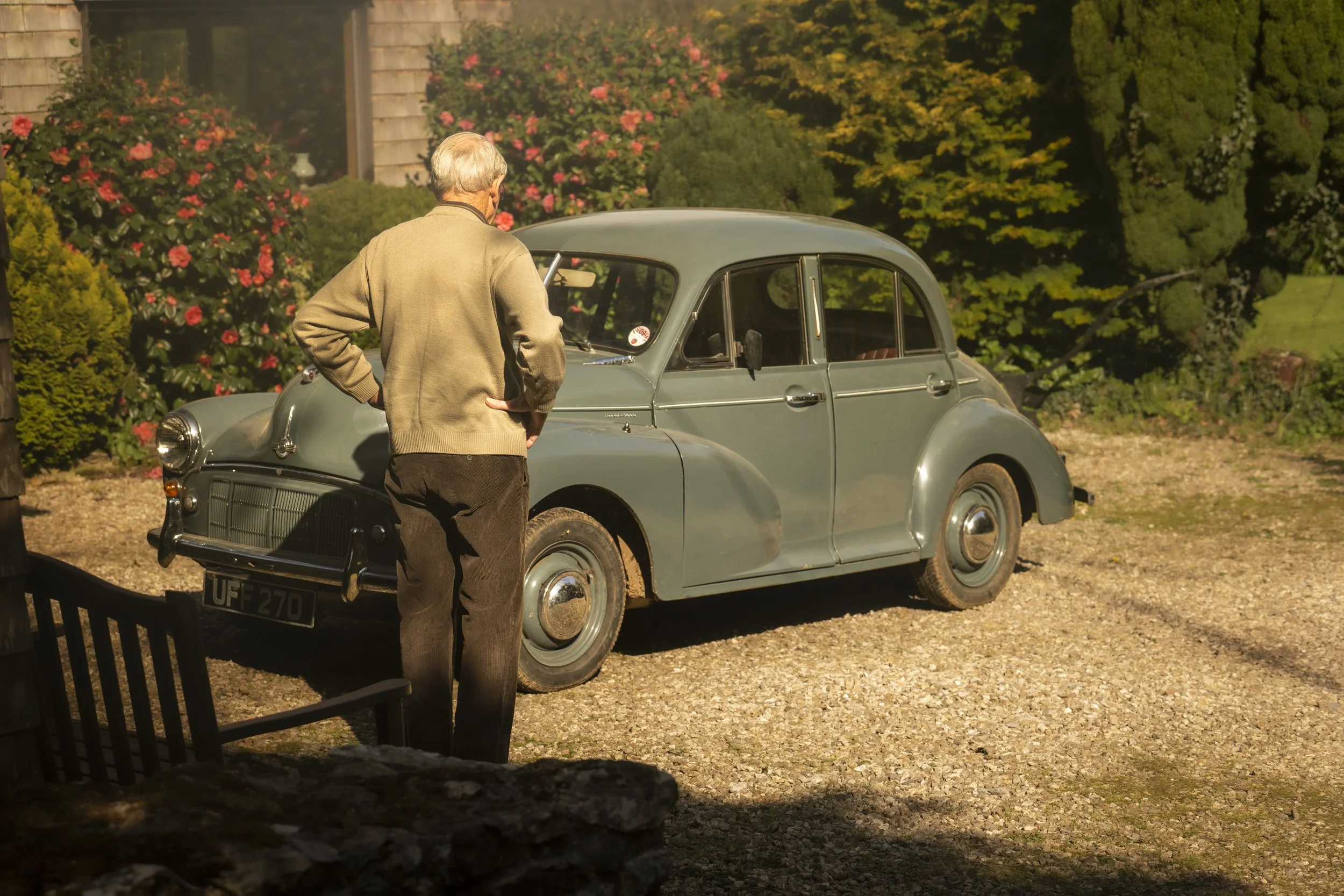 An older man with gray hair, wearing a beige cardigan and brown pants, stands outside looking at a vintage gray car, surrounded by colorful blooming bushes and trees.