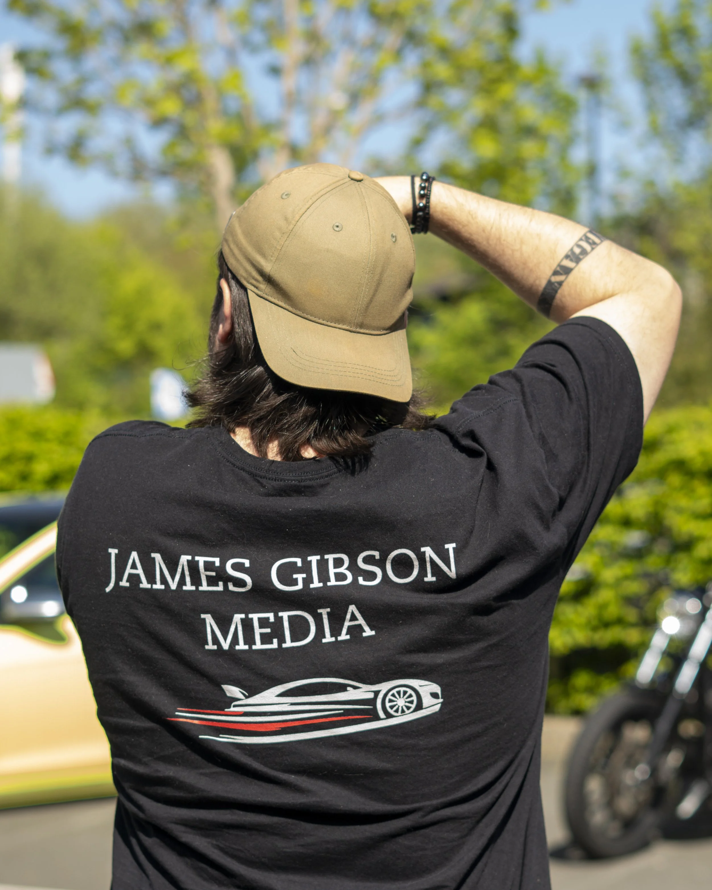 Person wearing a black T-shirt with white text that reads 'James Gibson Media' and a car graphic, with their back turned, outdoors with green trees and a blue sky in the background.