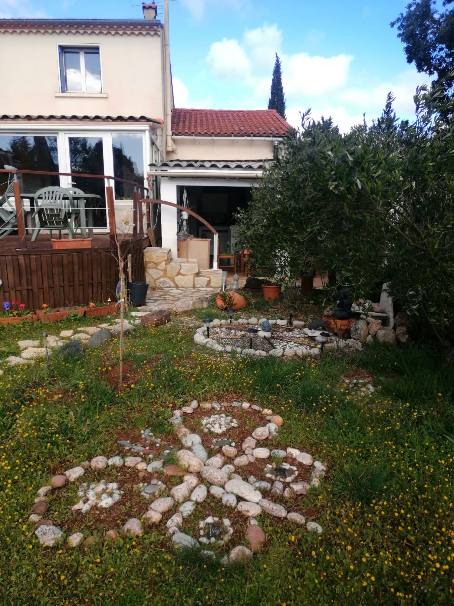 Jardin avec plantes, maison en arrière-plan, terrasse en bois, ornements en pierre et pelouse verte.