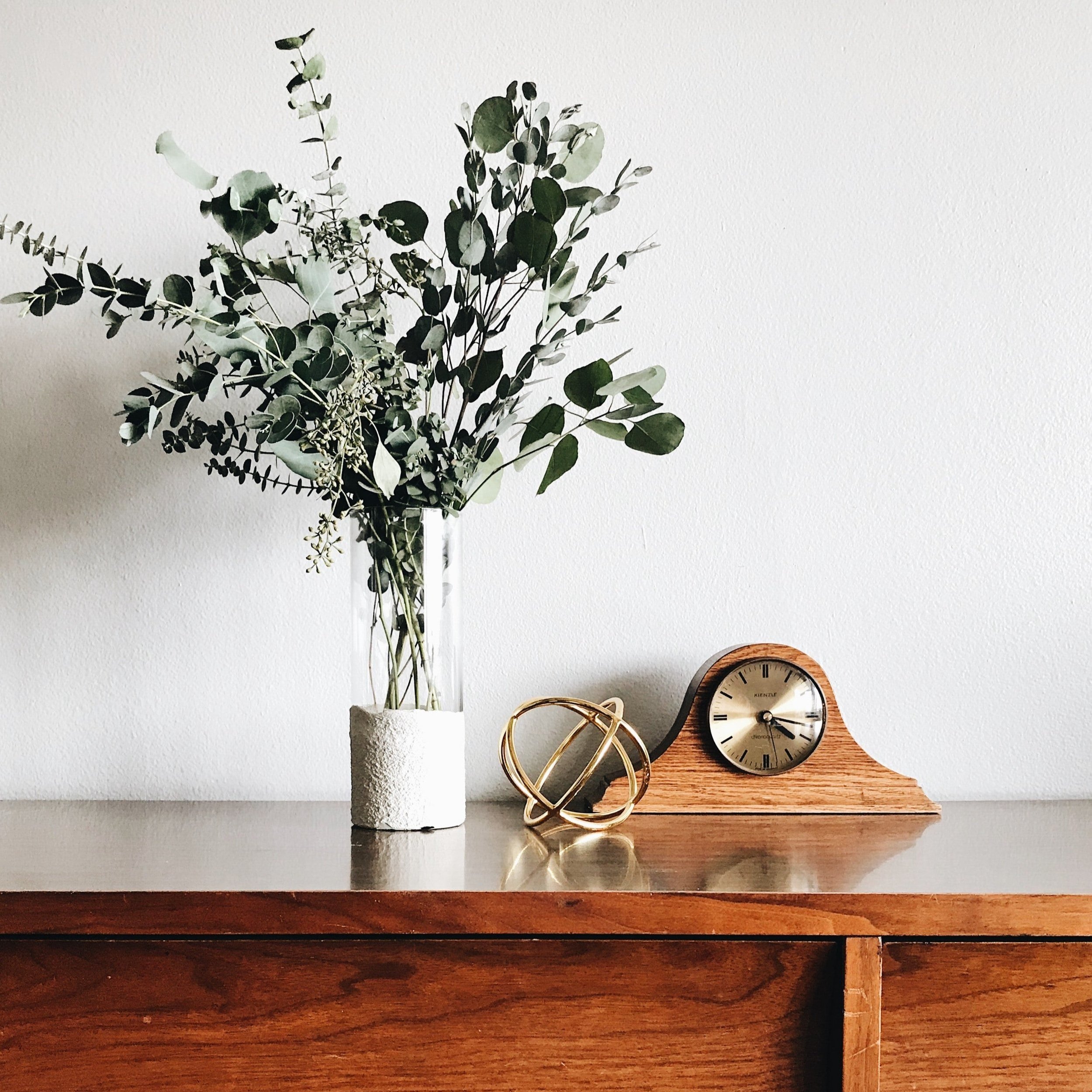 Grands feuillages verts dans un vase en verre sur un meuble en bois, à côté d'une petite horloge en bois et d'un objet décoratif doré.