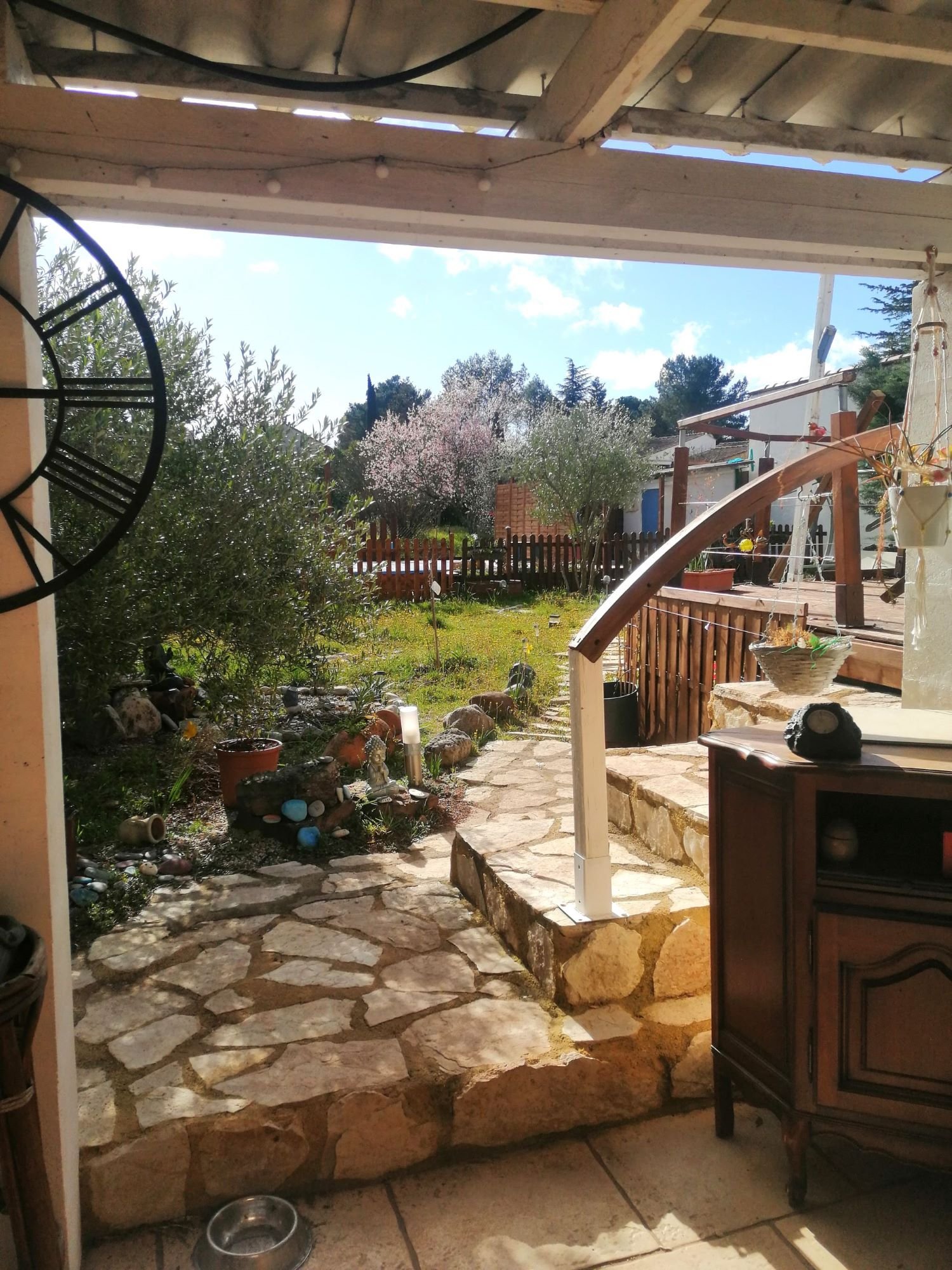 Jardin avec terrasse en pierre vue depuis une véranda. Arbres en fleurs et ciel bleu. Mobilier de jardin en bois et clôture en bois aussi visible. Pot de fleurs et décorations extérieures.