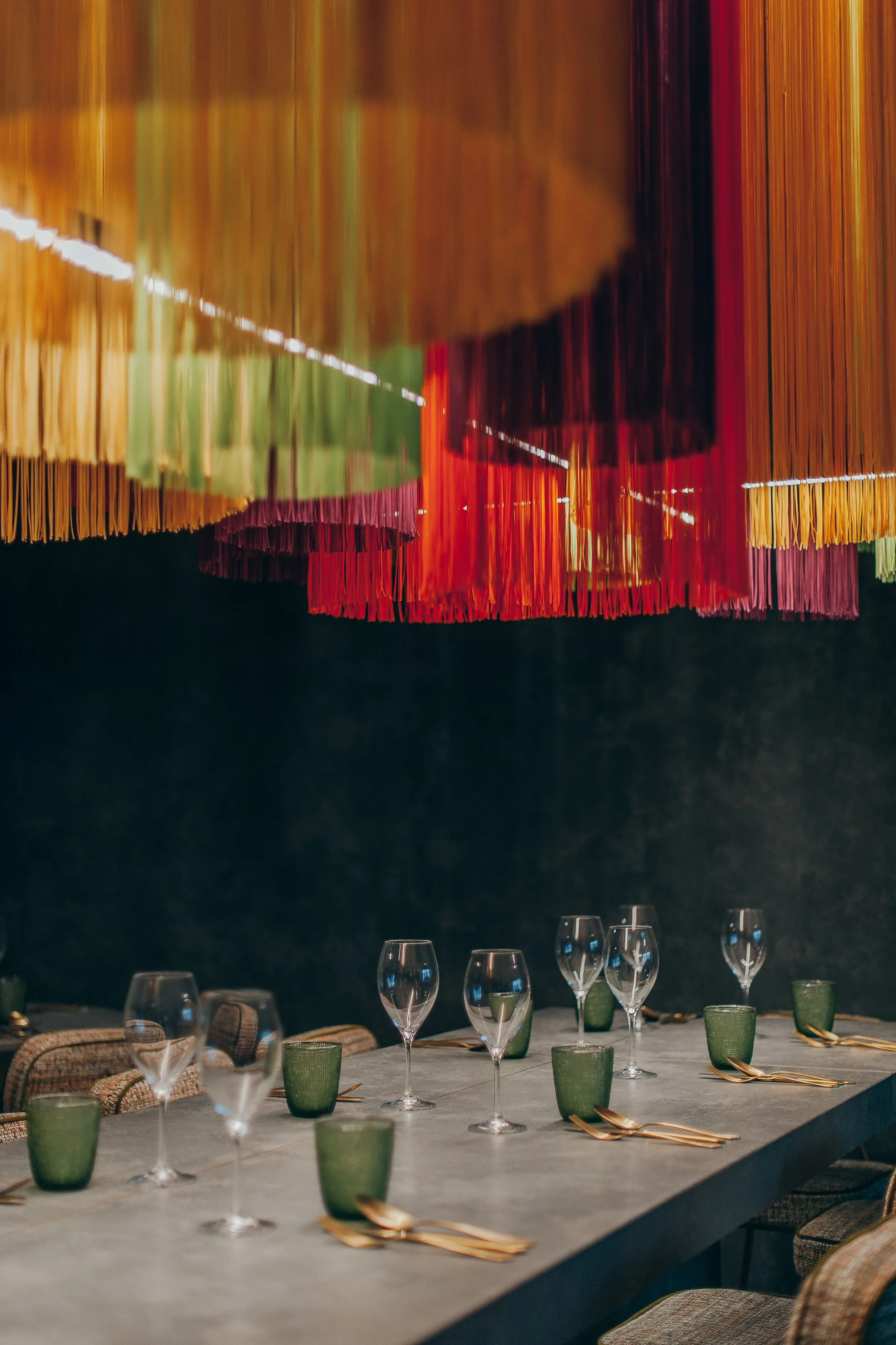 Table set with wine glasses, green cups, and gold utensils, with colorful hanging fabric decorations overhead in a dimly lit room.