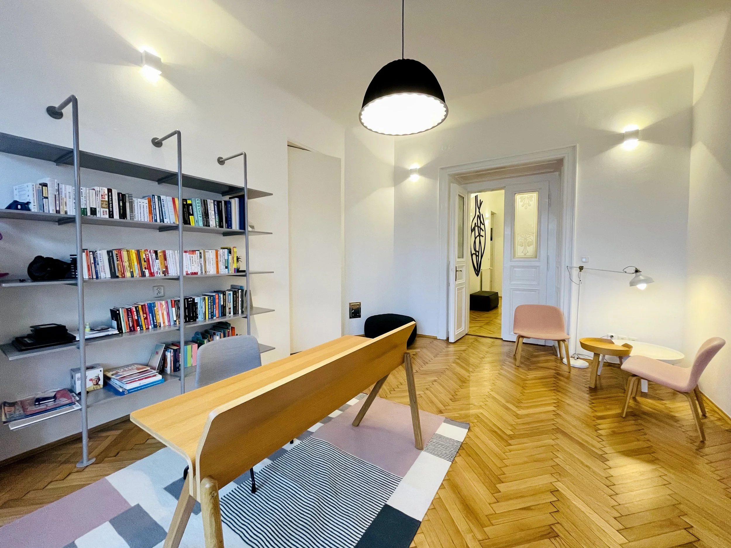 Interior of a living room with wooden floor, a large shelf filled with books, a wooden table, pink chairs, and unique lighting fixtures.
