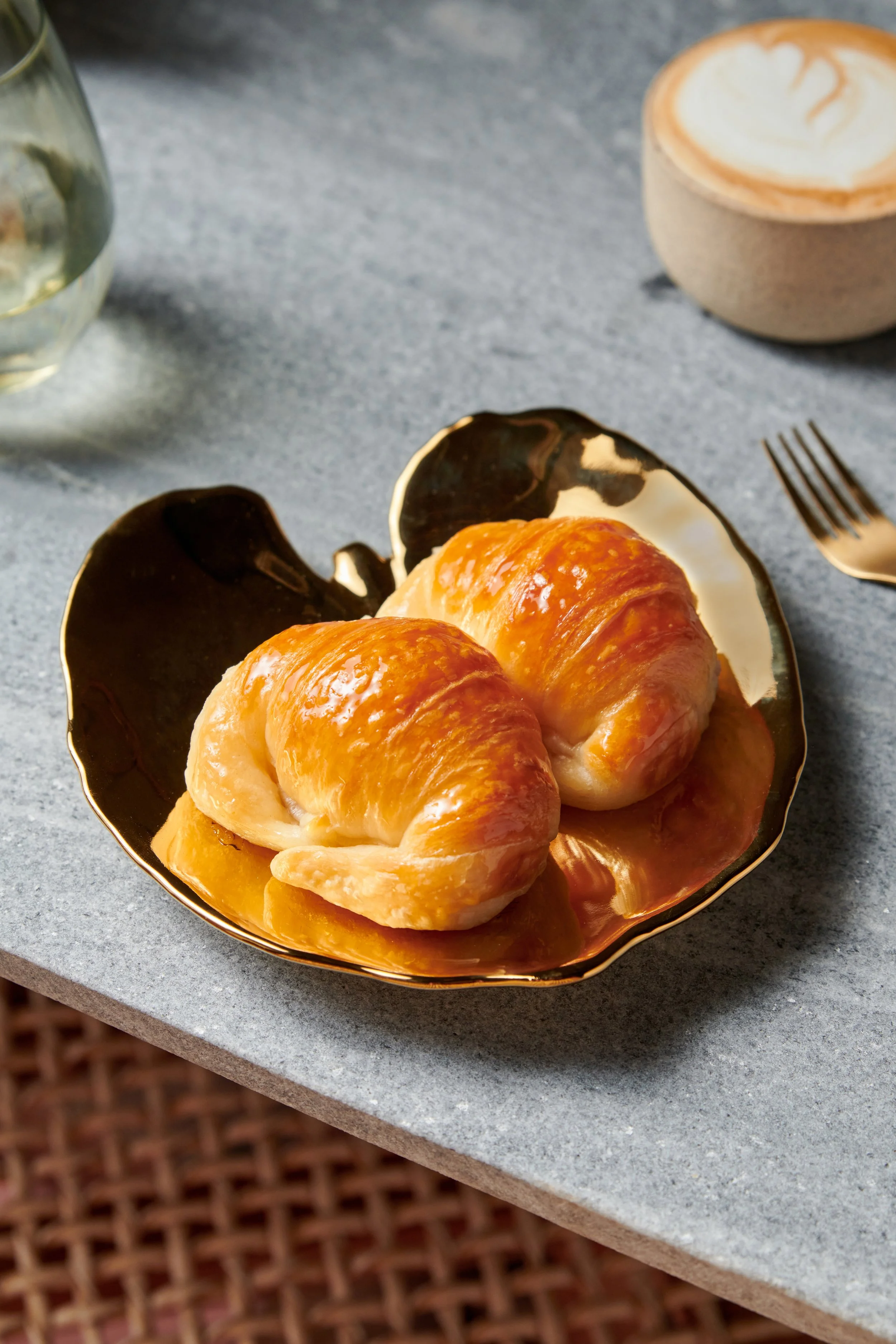 Two croissants on a gold plate placed on a gray table, with a glass of water, a latte in a horn-shaped cup, and a fork nearby.