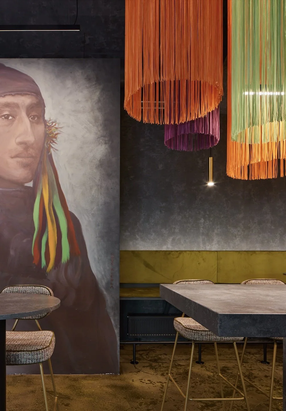 Interior of a modern restaurant with a portrait of a man with colorful dreadlocks on the wall, vibrant hanging fabric chandeliers in orange, purple, and green, a concrete table, and contemporary chairs.