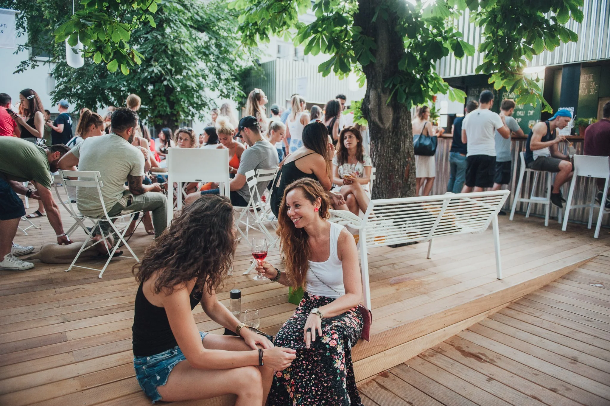 Two women sitting on a wooden deck, one with long curly hair in a black tank top and denim shorts, the other with long straight hair and a floral skirt, enjoying a glass of red wine and chatting amidst a lively outdoor gathering with people sitting, standing, and walking around, trees and a bar in the background.