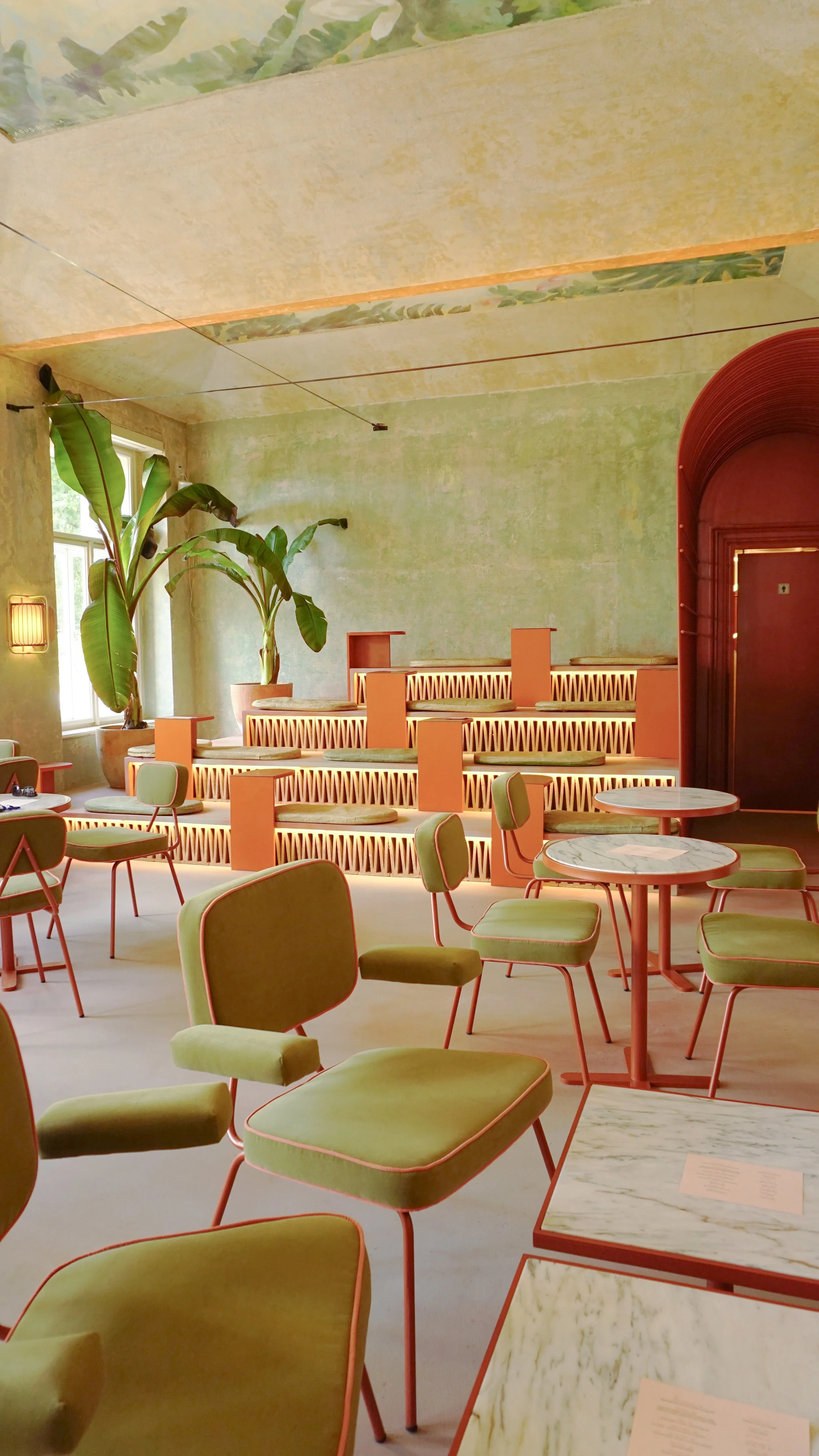 Interior of a modern cafe with green chairs, marble tables, large potted plants, and tiered seating with pink accents and lighting.