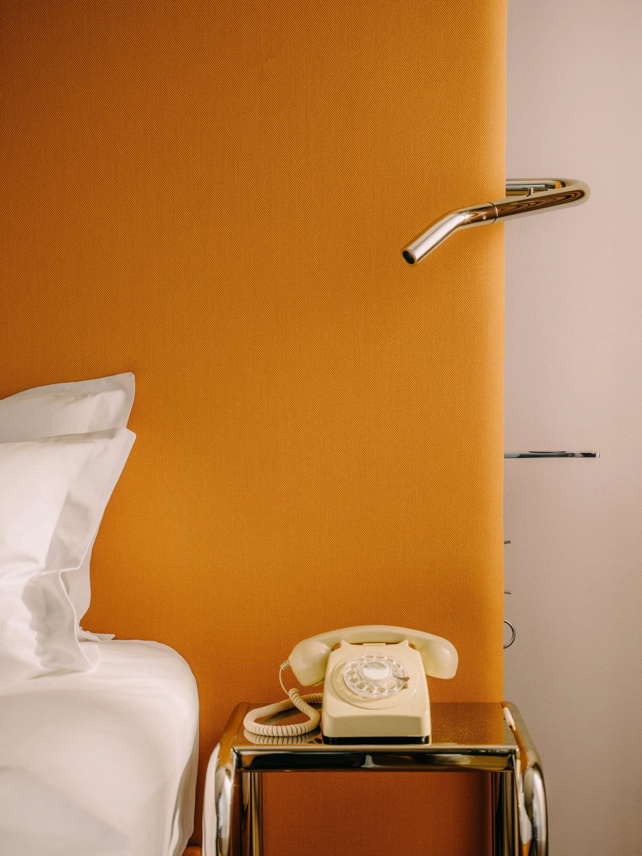A bedside table with a vintage rotary phone and part of a bed with white sheets and pillow, against an orange headboard with a chrome reading lamp attached.