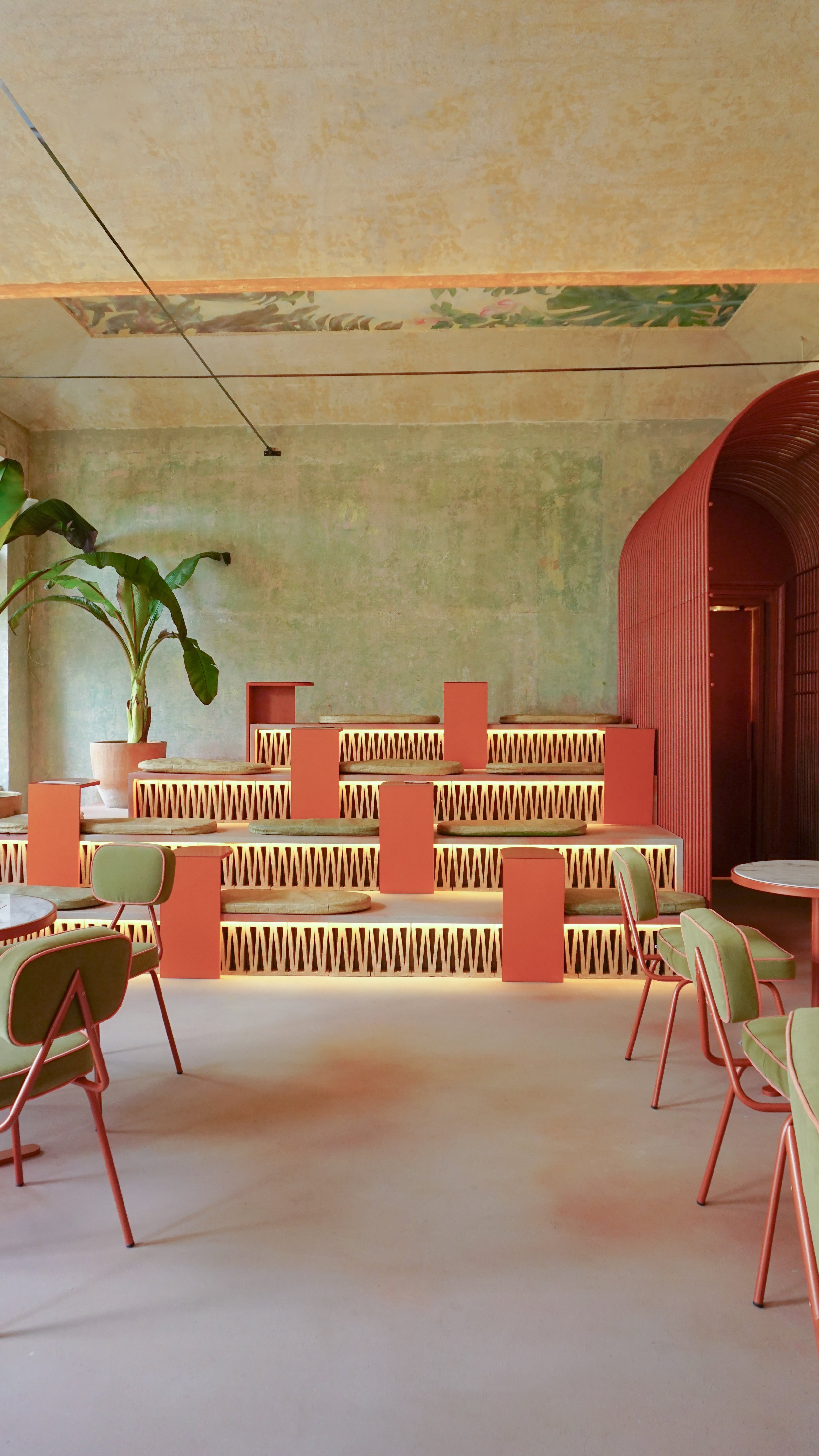 Modern indoor cafe with tiered seating, pink and green decor, chairs with pink frames and green cushions, potted large plant in the corner, and abstract wall art.