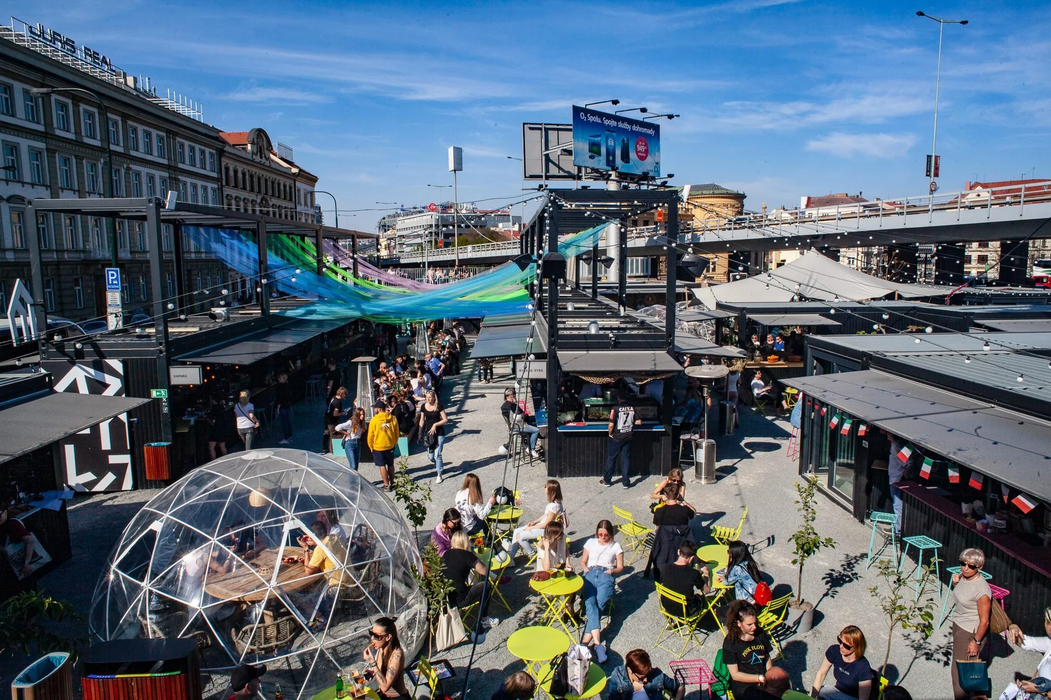 Outdoor market or food court with people sitting at colorful tables, standing, and walking. There are various vendor stalls, a geodesic dome structure, and decorative hanging lights under a clear blue sky.