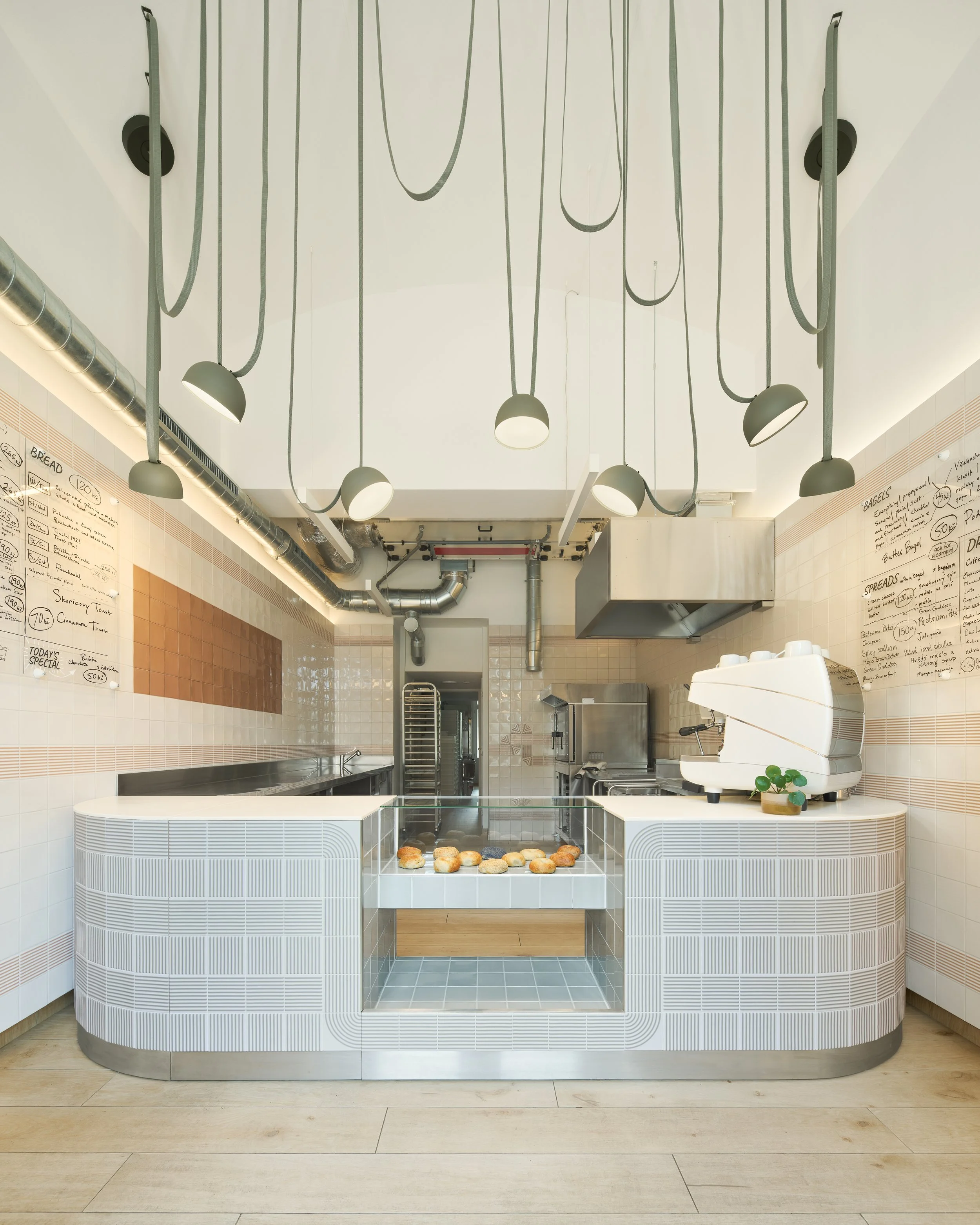 Interior of a modern bakery or cafe with a white display counter filled with bread rolls, and pendant lights hanging from the ceiling with cords. Menu boards with handwritten items are mounted on tiled walls on either side.