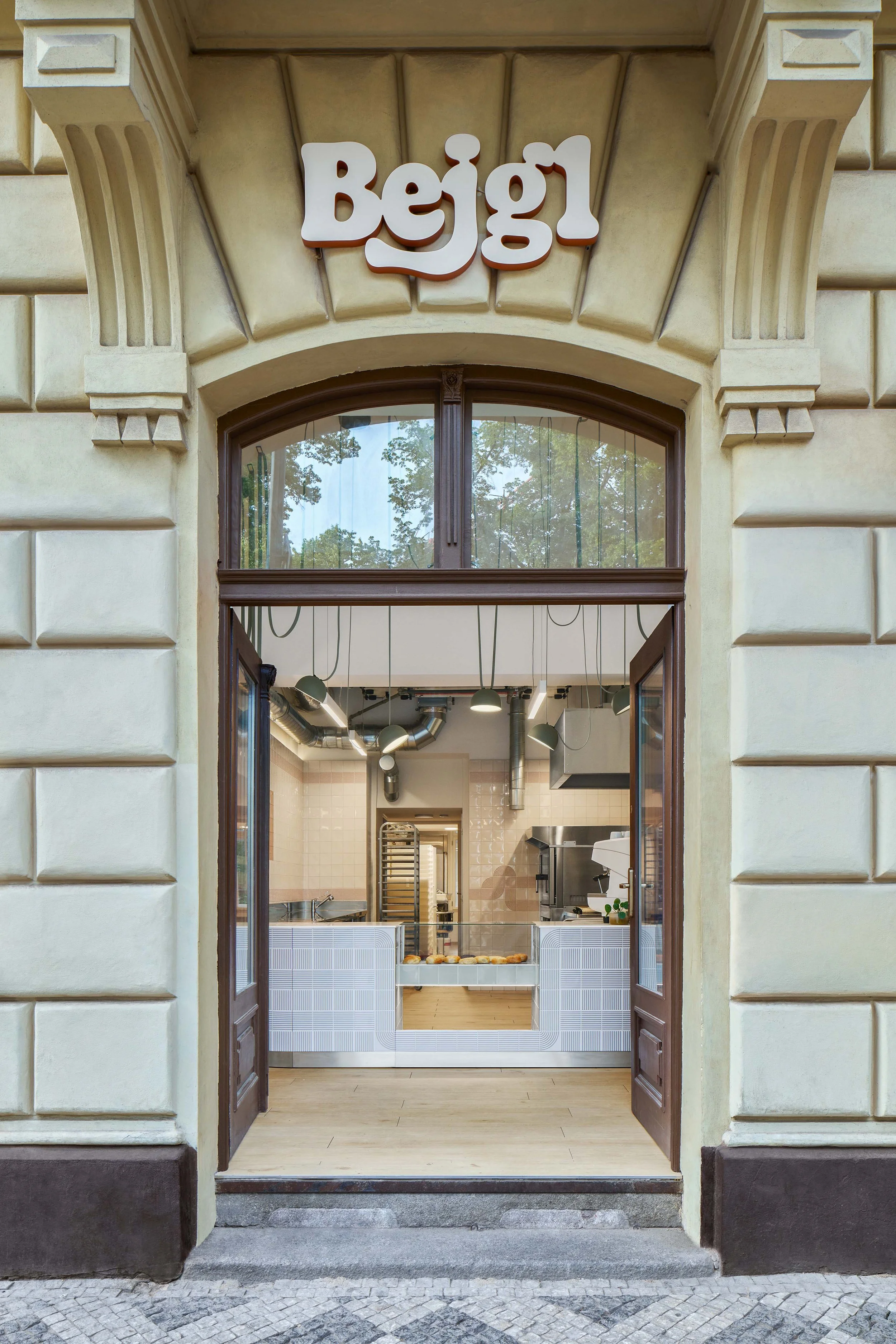 View of a bakery shop entrance with a sign that reads 'Beigel', showing the inside kitchen with baking trays and equipment.