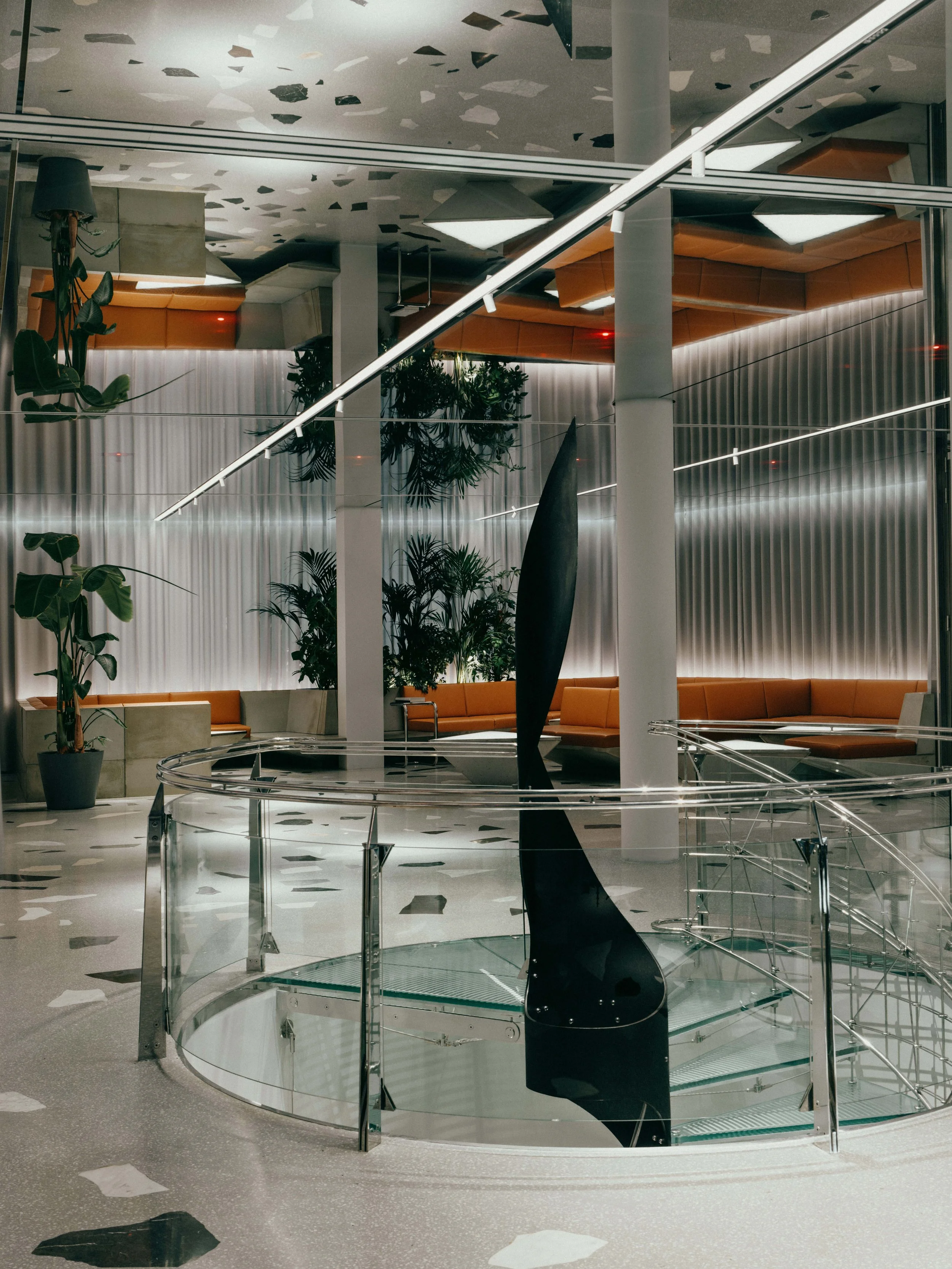 Modern interior space featuring a black sculptural art piece inside a glass-enclosed spiral staircase, with leafy green plants and orange seating areas in the background.