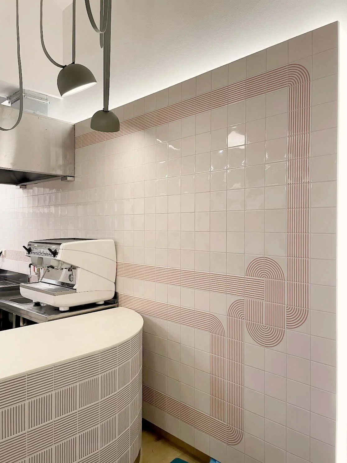 Interior of a cafe or restaurant kitchen with a tiled wall, a coffee machine on a counter, and modern overhead lighting.