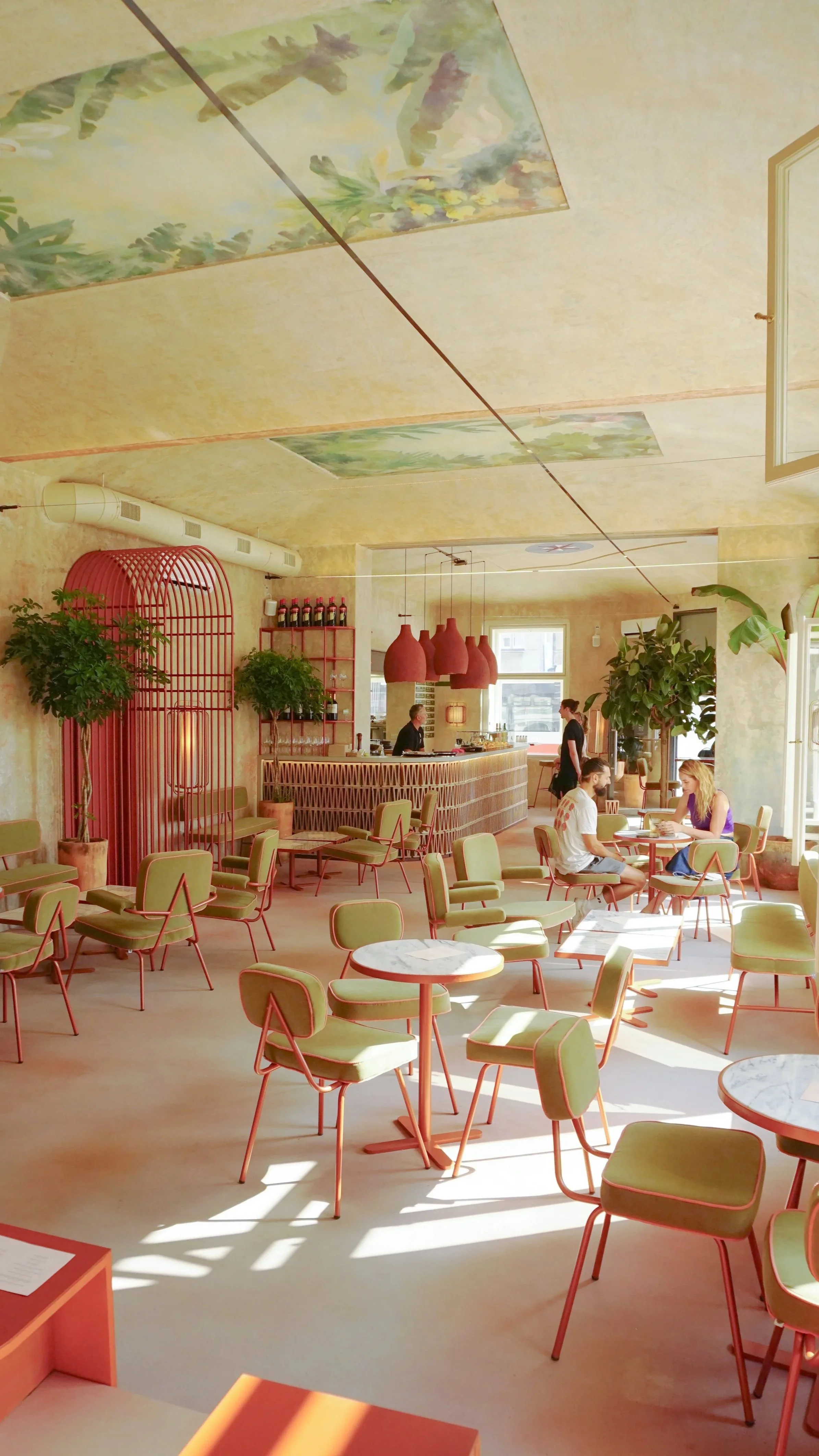 A bright, modern cafe with green chairs, marble-topped tables, potted plants, and a colorful ceiling mural. There are two people sitting at a table near the window, and staff behind the counter with three large red pendant lights hanging above.
