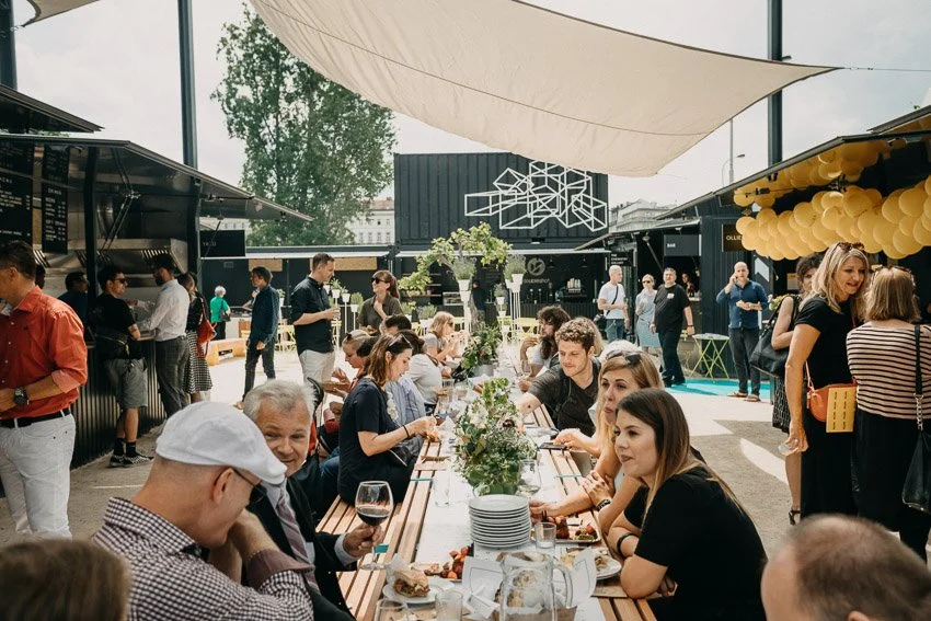People dining at long table during outdoor event with food, drinks, and floral centerpieces under large shade sail in a modern outdoor setting.