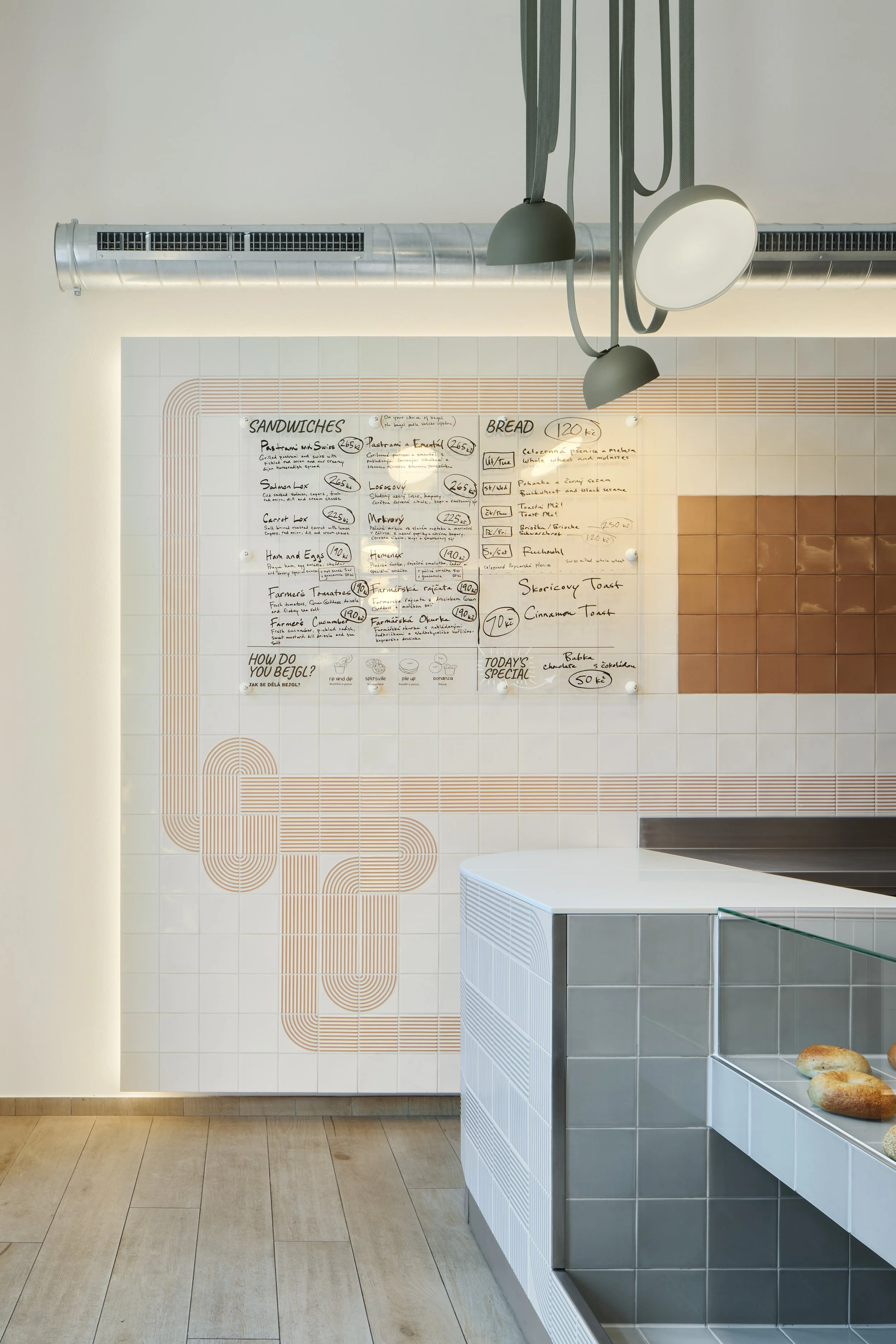 A modern bakery interior with a white tiled counter and bagels on it, a whiteboard menu on the wall with handwritten items and minimalistic lighting fixtures.