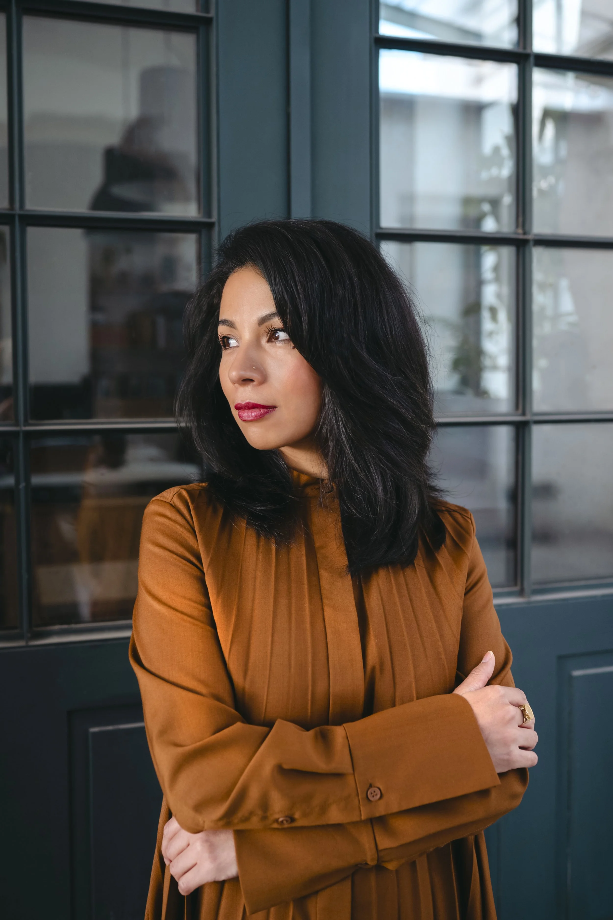 A woman with black hair and a nose piercing, wearing a brown long-sleeved dress, stands in front of a dark green windowed door, looking to her right with arms crossed.