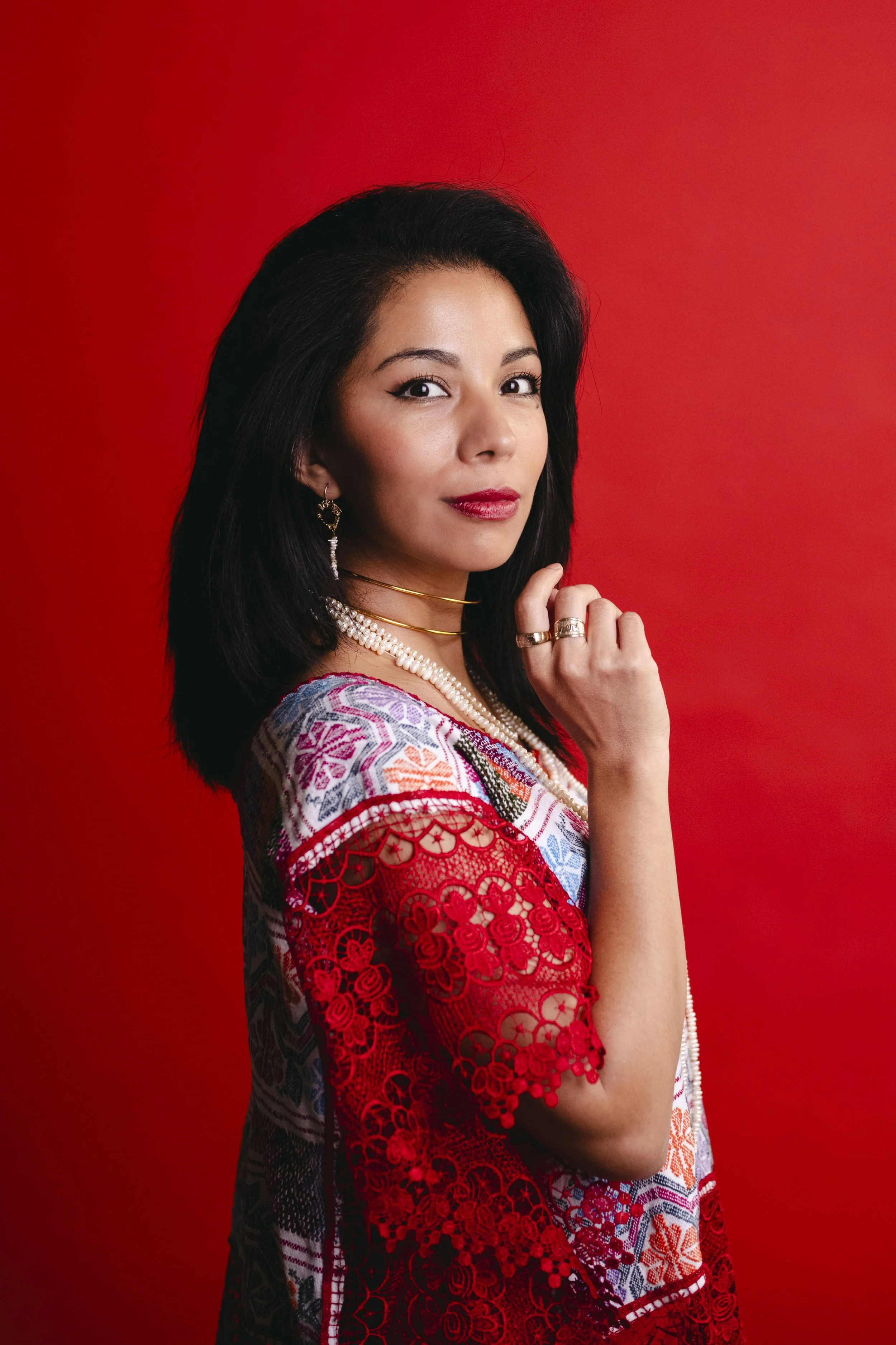 A woman with dark hair, red lipstick, and jewelry, wearing a colorful embroidered dress with red lace details, stands against a red background, gazing confidently at the camera.