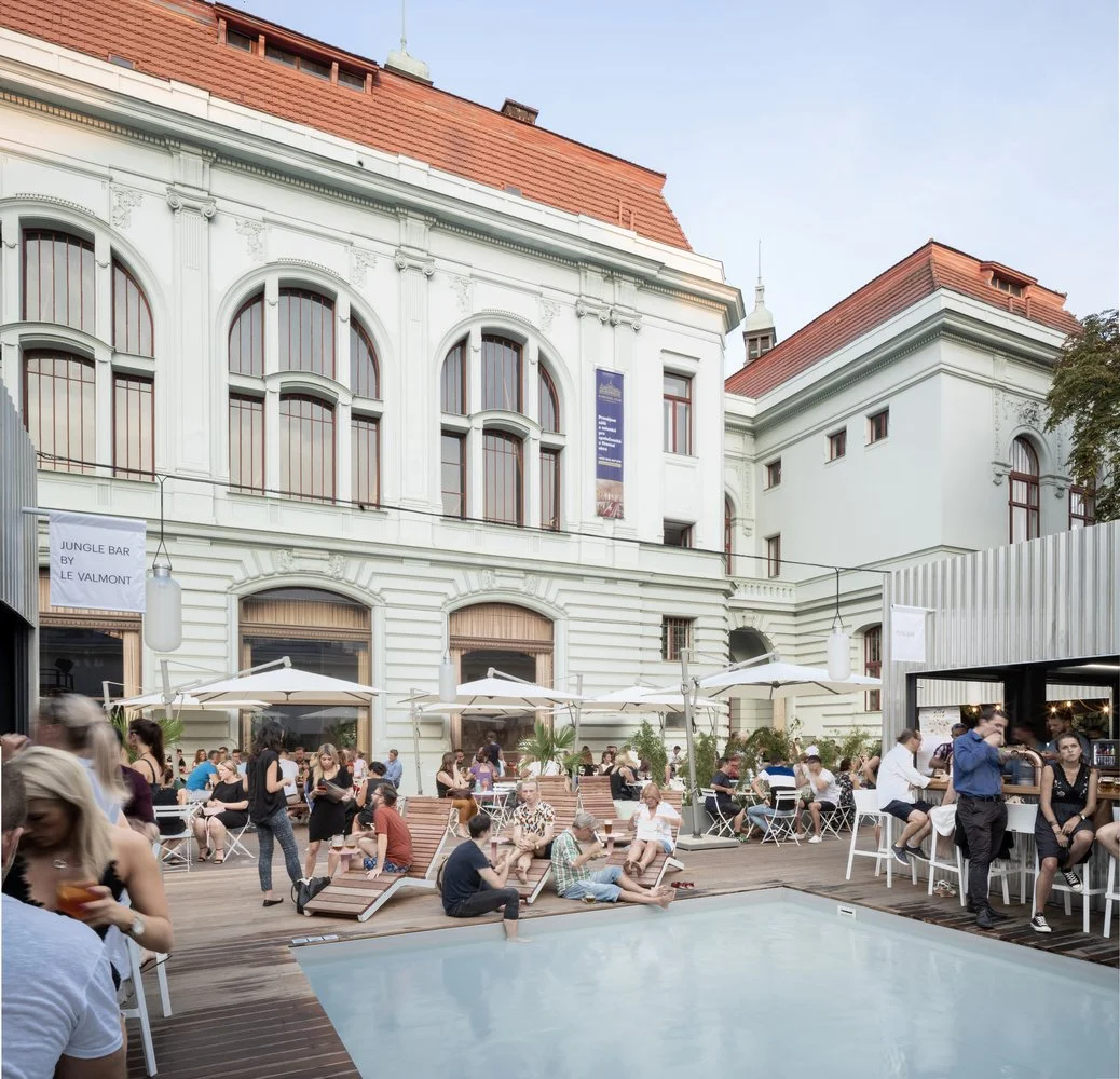 People relaxing and socializing on a rooftop terrace with a pool, surrounded by white historic buildings, umbrellas, and lounge chairs.