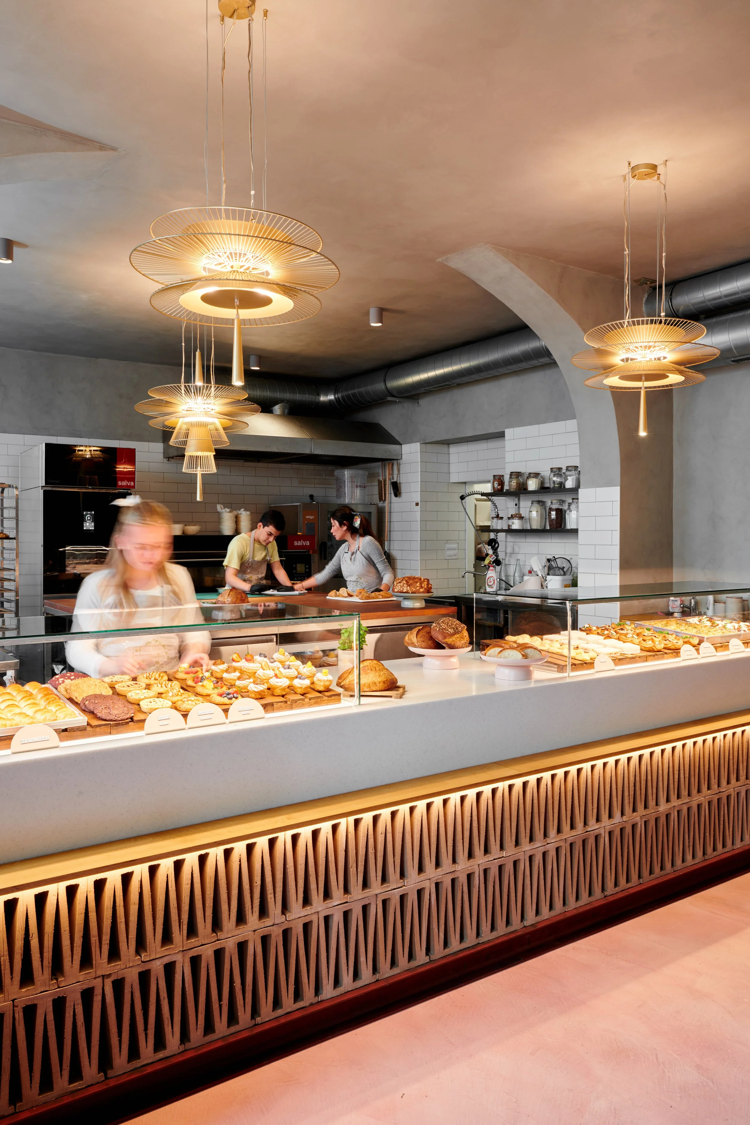 A bakery display case filled with various cookies, pastries, and breads in a modern bakery or cafe. In the background, two staff members prepare baked goods behind the counter, with additional baked items displayed on top of the case. The cafe has stylish lighting and a contemporary interior.