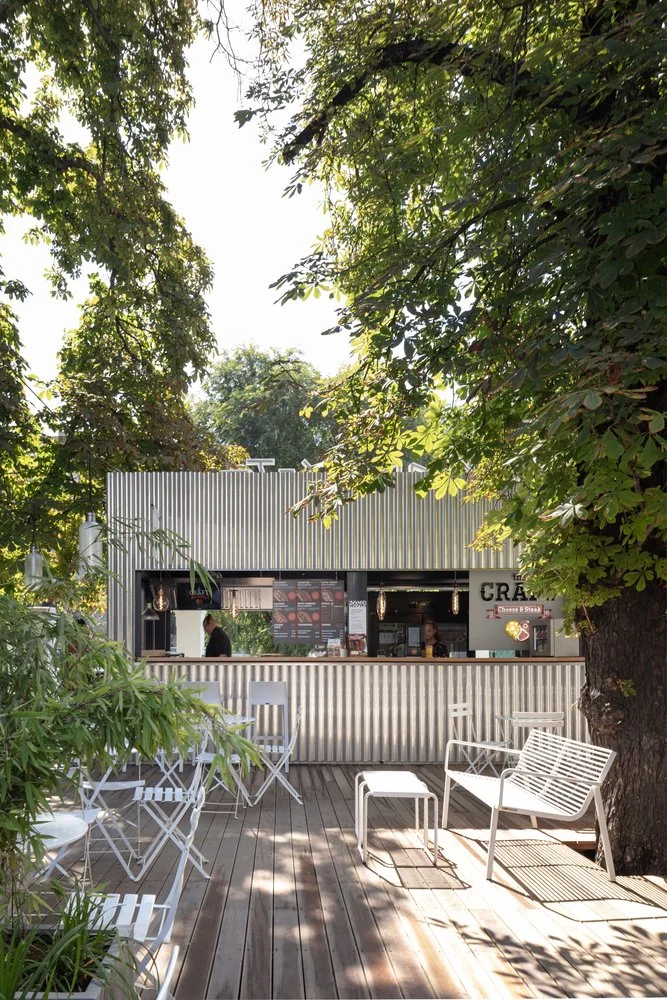 Outdoor food stand with a corrugated metal facade, business sign, and a menu, surrounded by trees and seating with white chairs and benches on a wooden deck