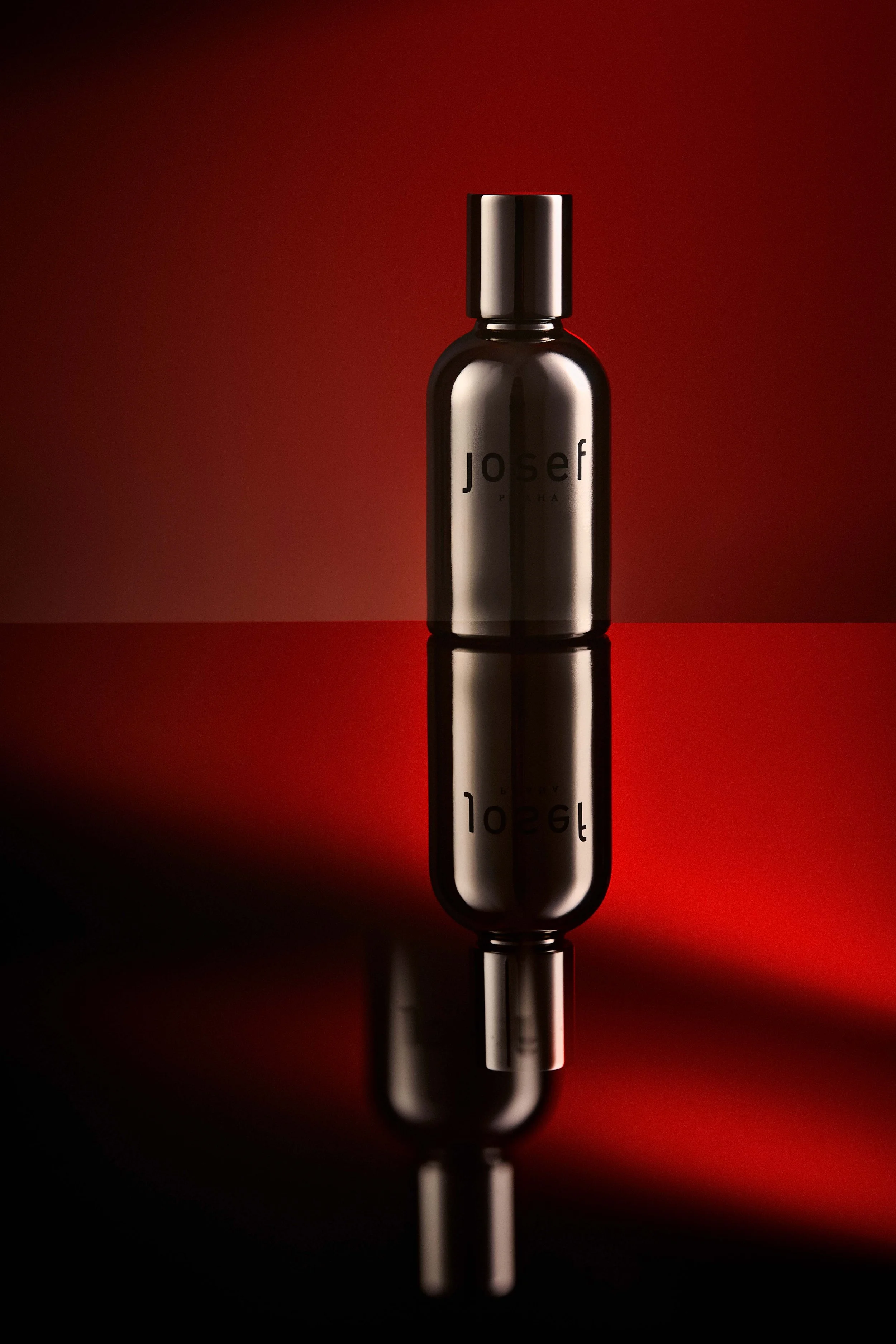 A reflective black bottle with the brand name 'Josef' on it, standing upright on a mirror surface with a dark red background.