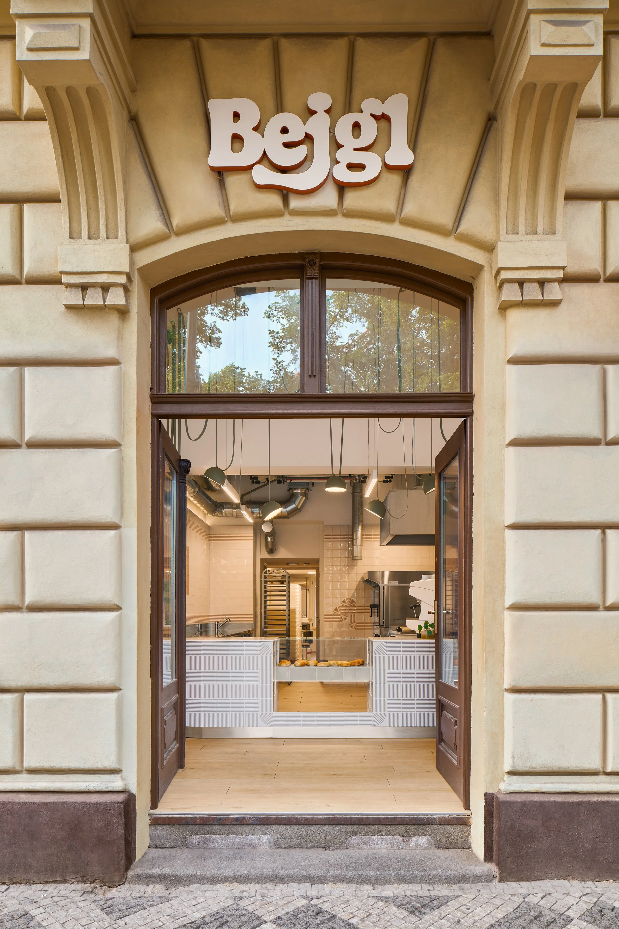 Bagel shop in Prague with a sign above the entrance that says 'Begl". The interior has a stainless-steel counter, tiles, and baked goods on display.