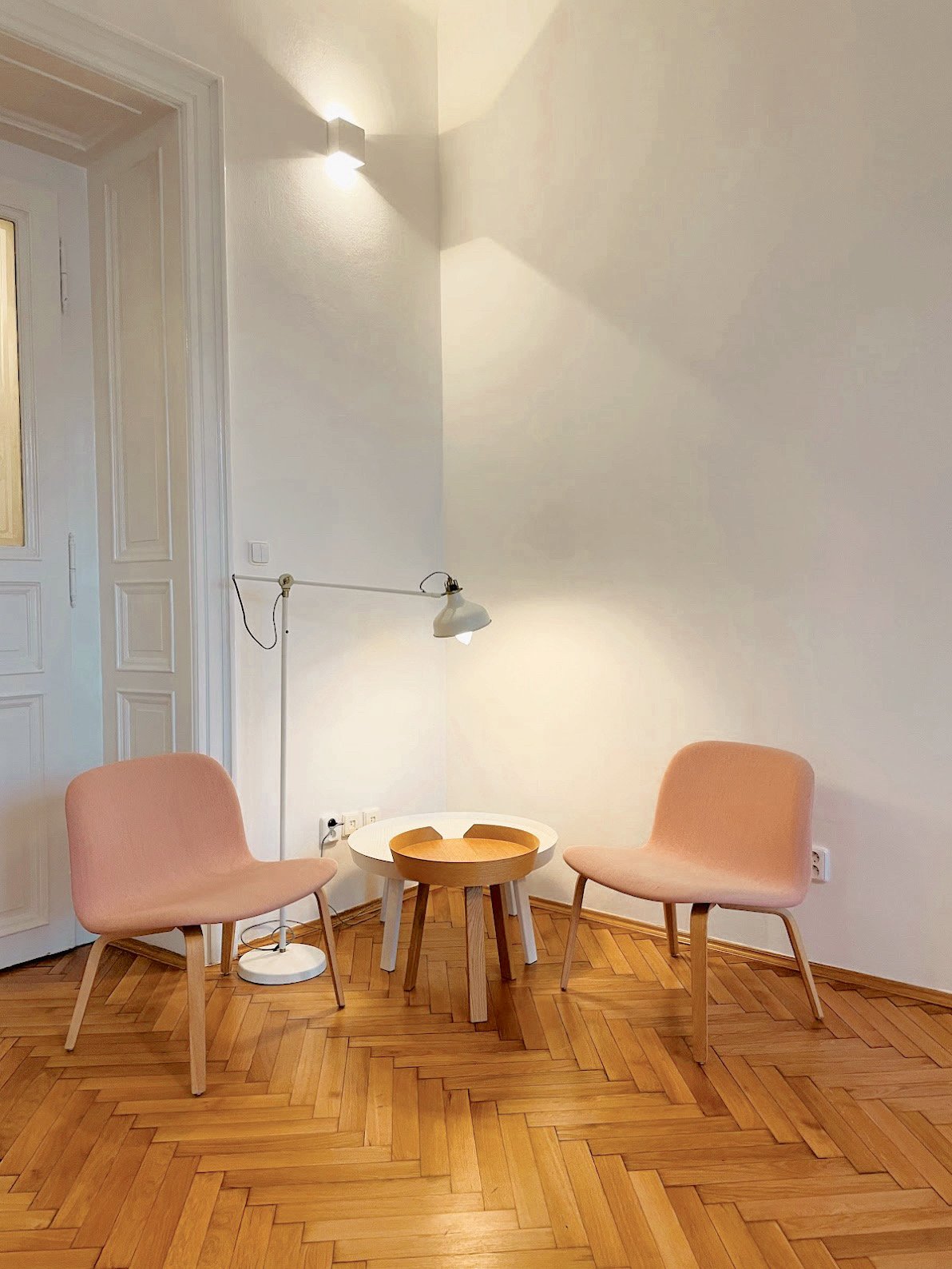A minimalist sitting area with two pink upholstered chairs, a small round wooden table, and a white adjustable floor lamp on a wooden parquet floor against a plain white wall.