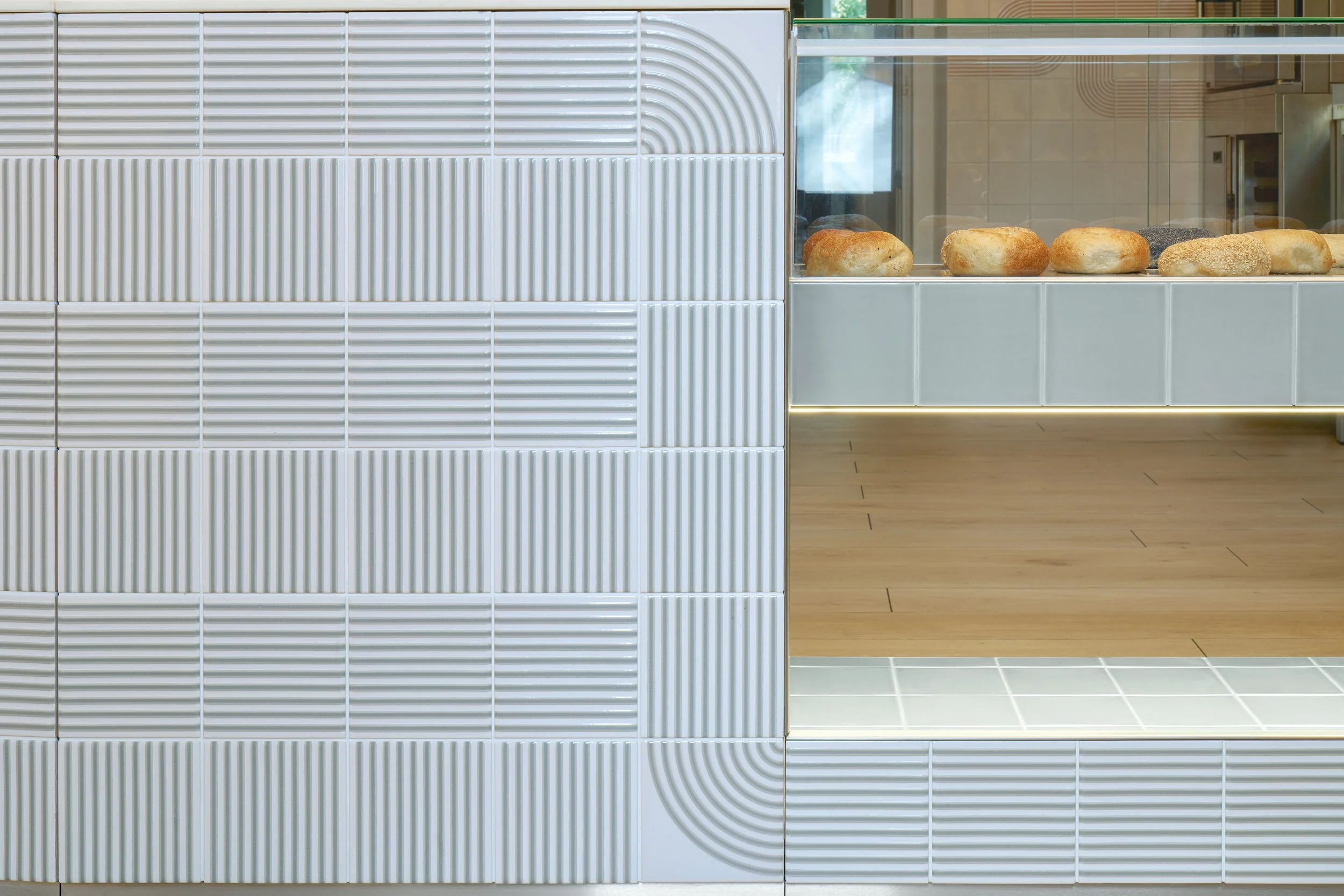 Display of assorted bread rolls inside a bakery display case with a modern, tiled wall behind.