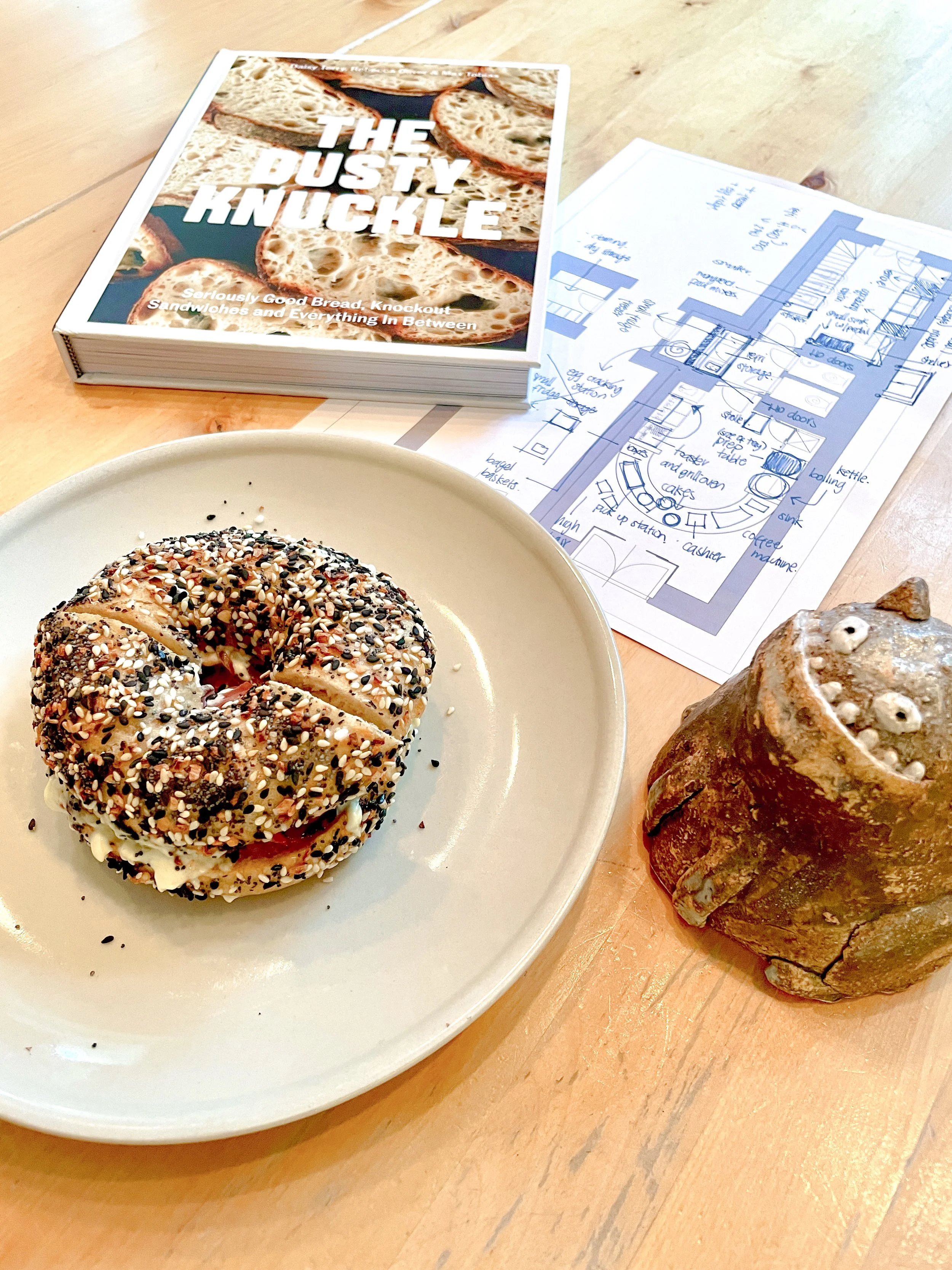 A bagel sandwich topped with sesame and black seeds on a white plate, a baked good with a swirl shape and white icing with small white and dark sprinkles on the side of the table, a book titled "The Dusty Knuckle" with a bread loaf on the cover, and a blue-sketch floor plan drawing of a restaurant interior.