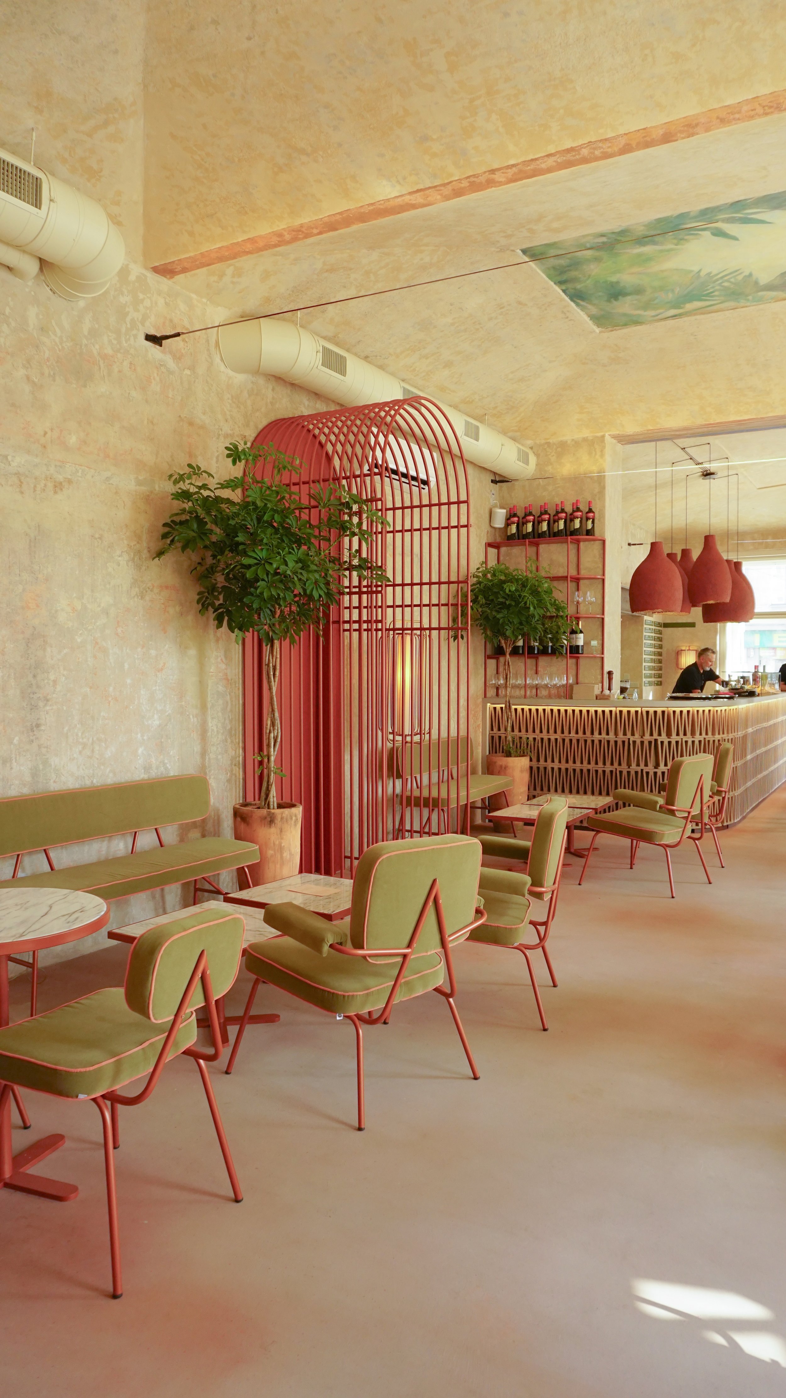 Modern cafe interior with pastel yellow walls, green chairs with pink outlines, potted plants, a red decorative room divider, and a bar area with hanging red pendant lights.