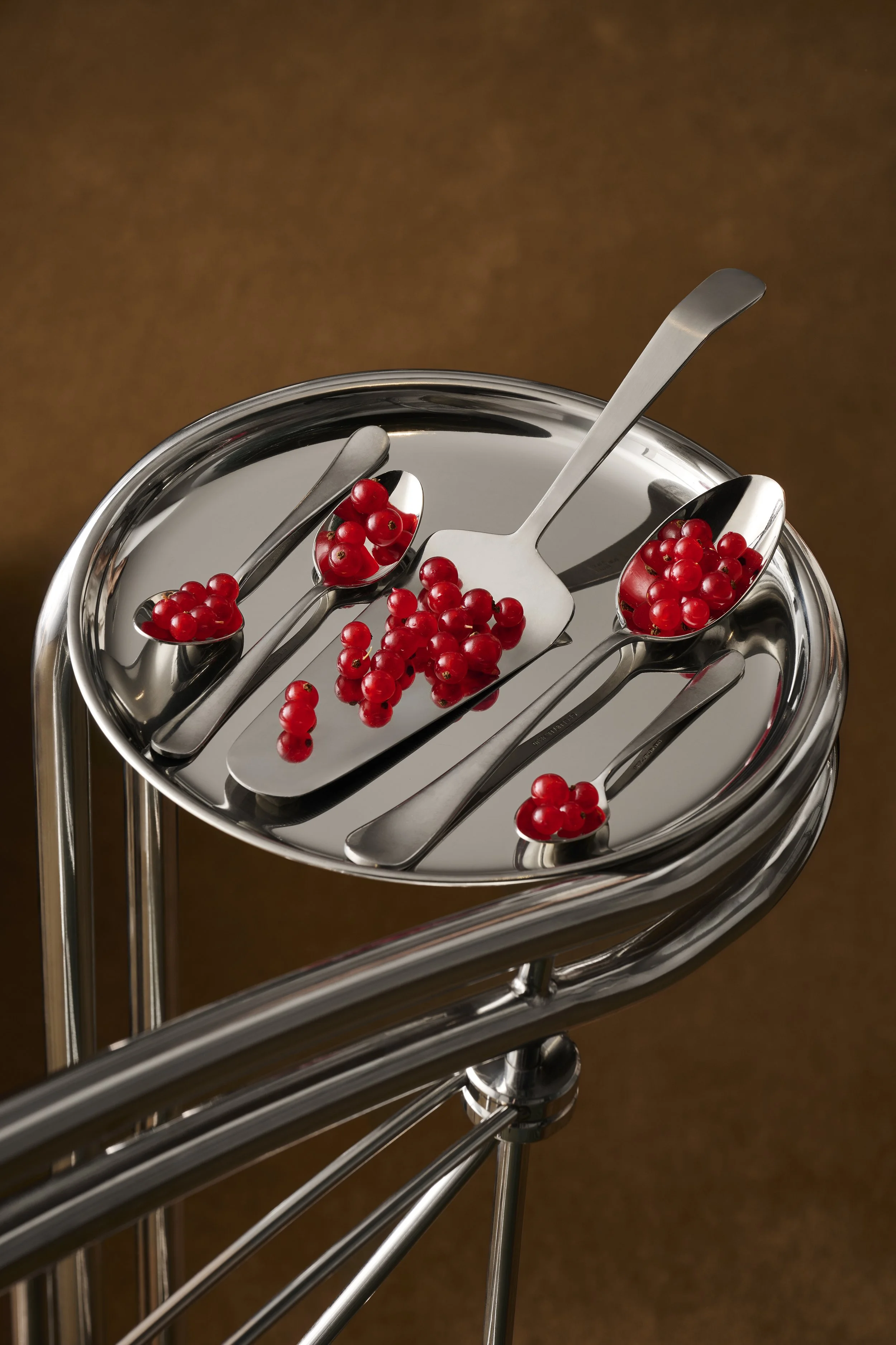 Red currants on stainless steel spoons and a scoop on a shiny metal serving cart with a brown background.