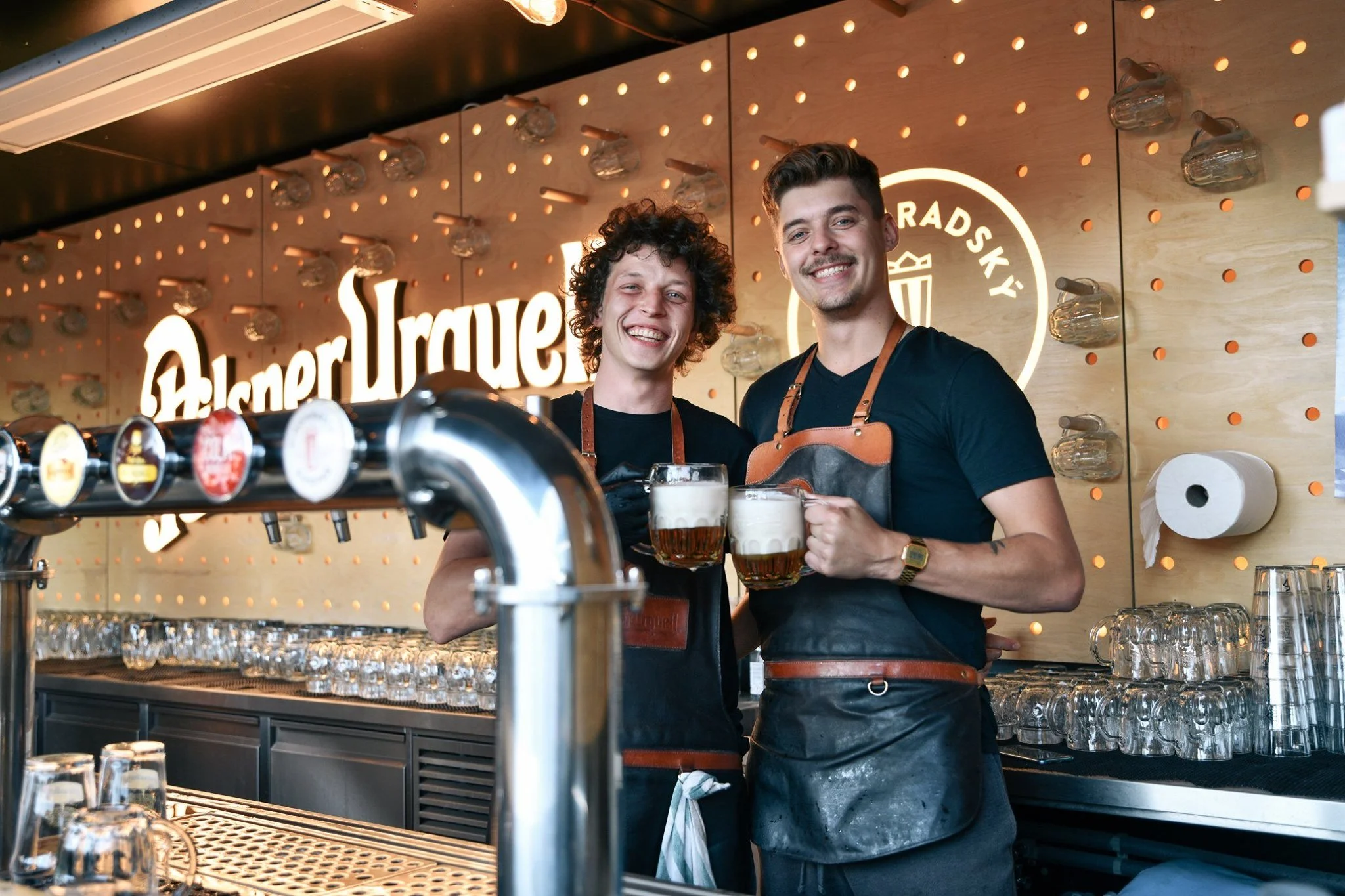 Two bartenders smiling and holding mugs of beer in front of a bar with beer taps, glasses, and a decorative wall with illuminated signage and string lights.