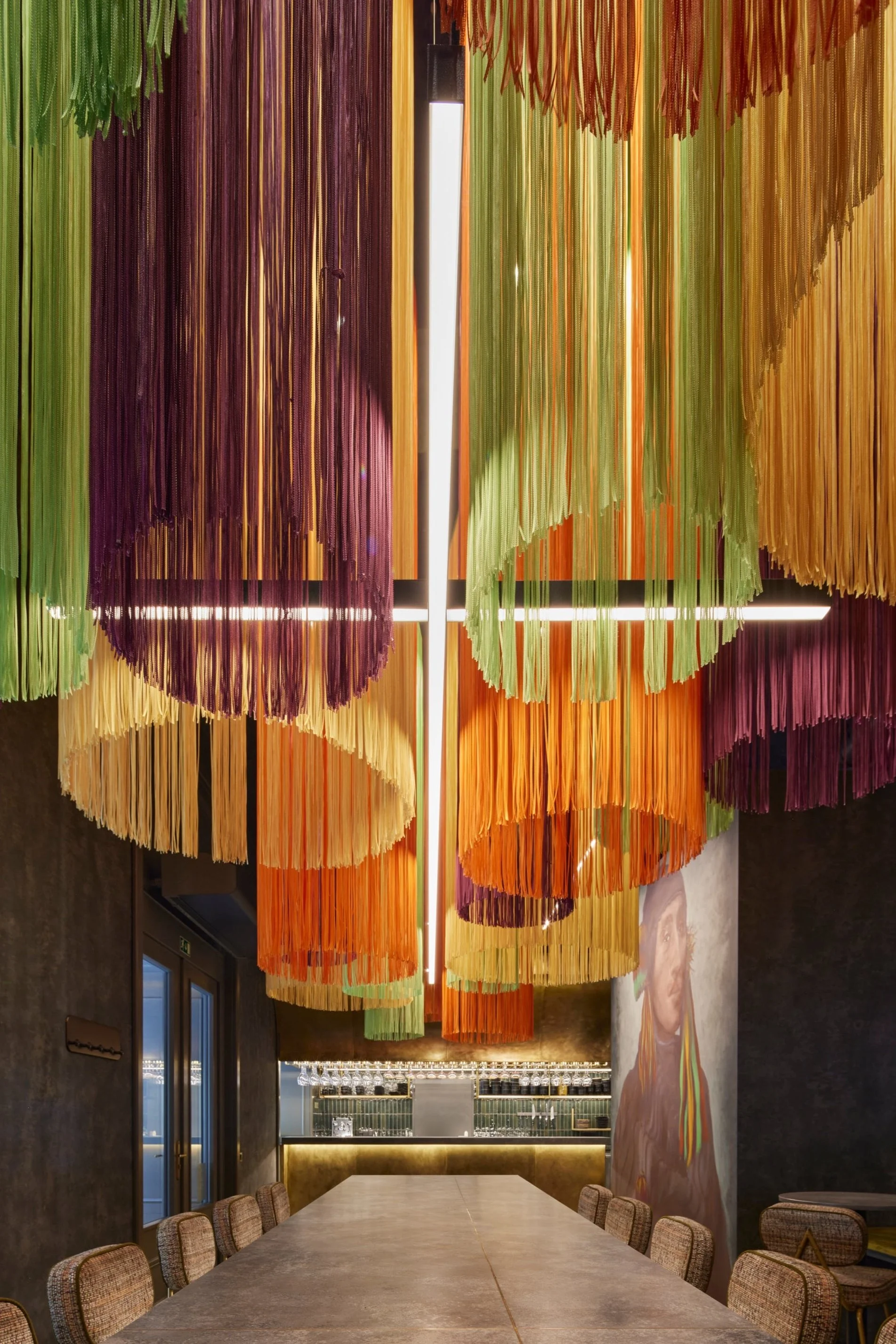 Colorful hanging art installation with strips of fabric or paper in purple, orange, green, and yellow, suspended from the ceiling in a modern restaurant or dining area.