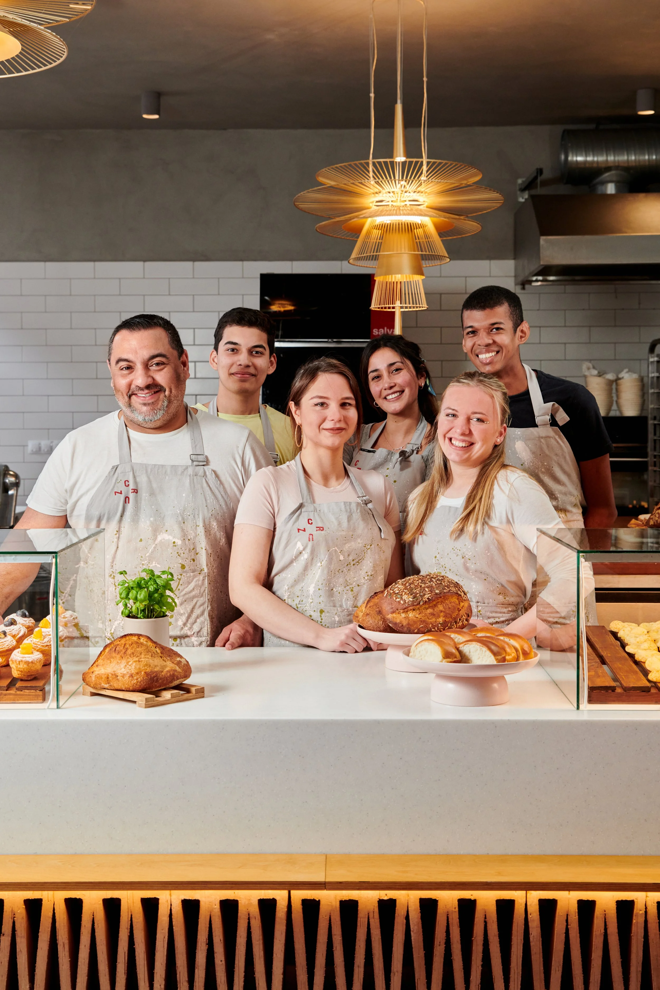 Six people standing behind a counter in a bakery, smiling, with bread and pastries on display, modern lighting and a white tiled wall in the background.