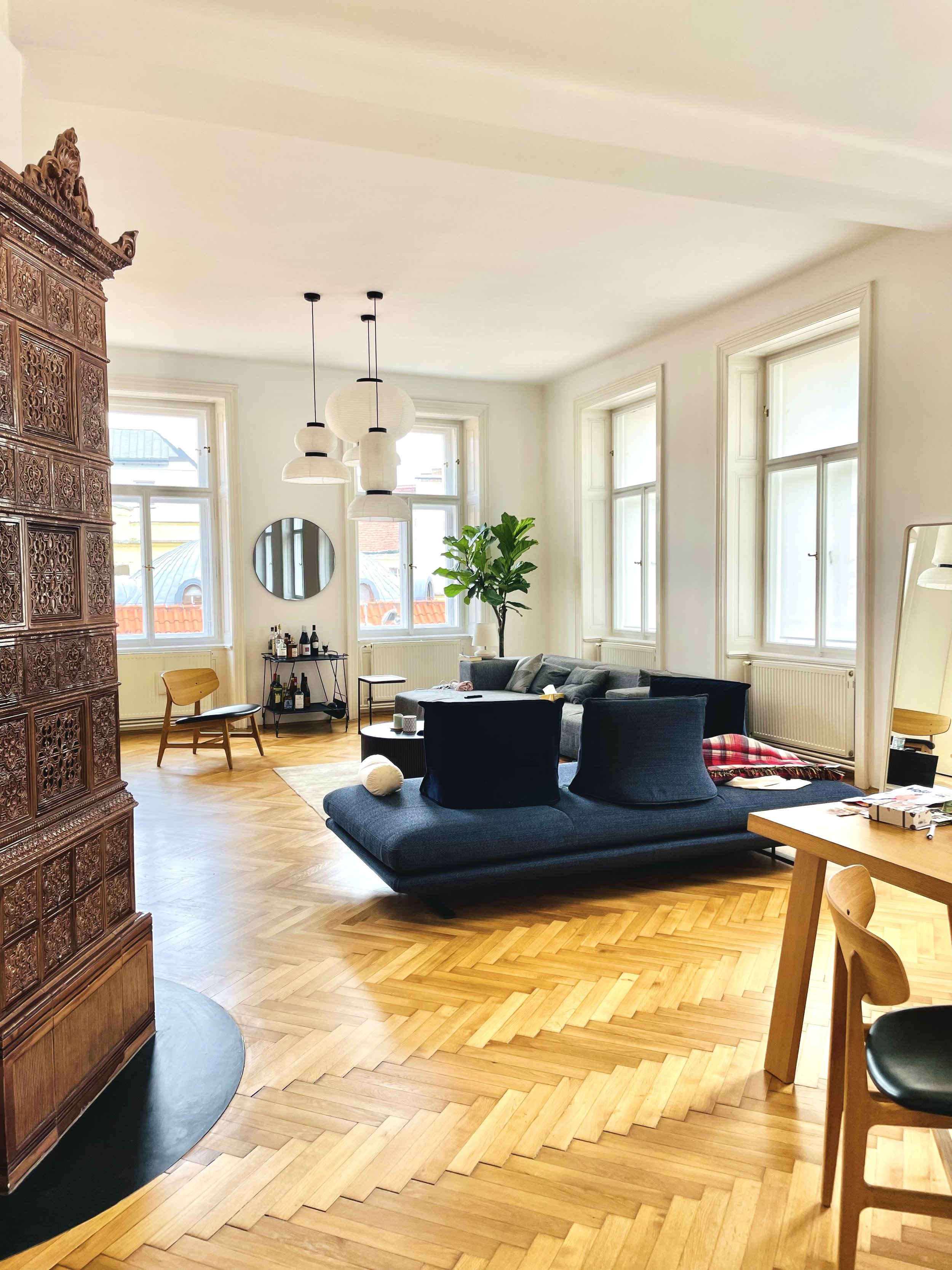 Bright living room with large windows, a gray sofa, a black lounge chair, a wooden dining table and chairs, a bar cart, and decorative items.