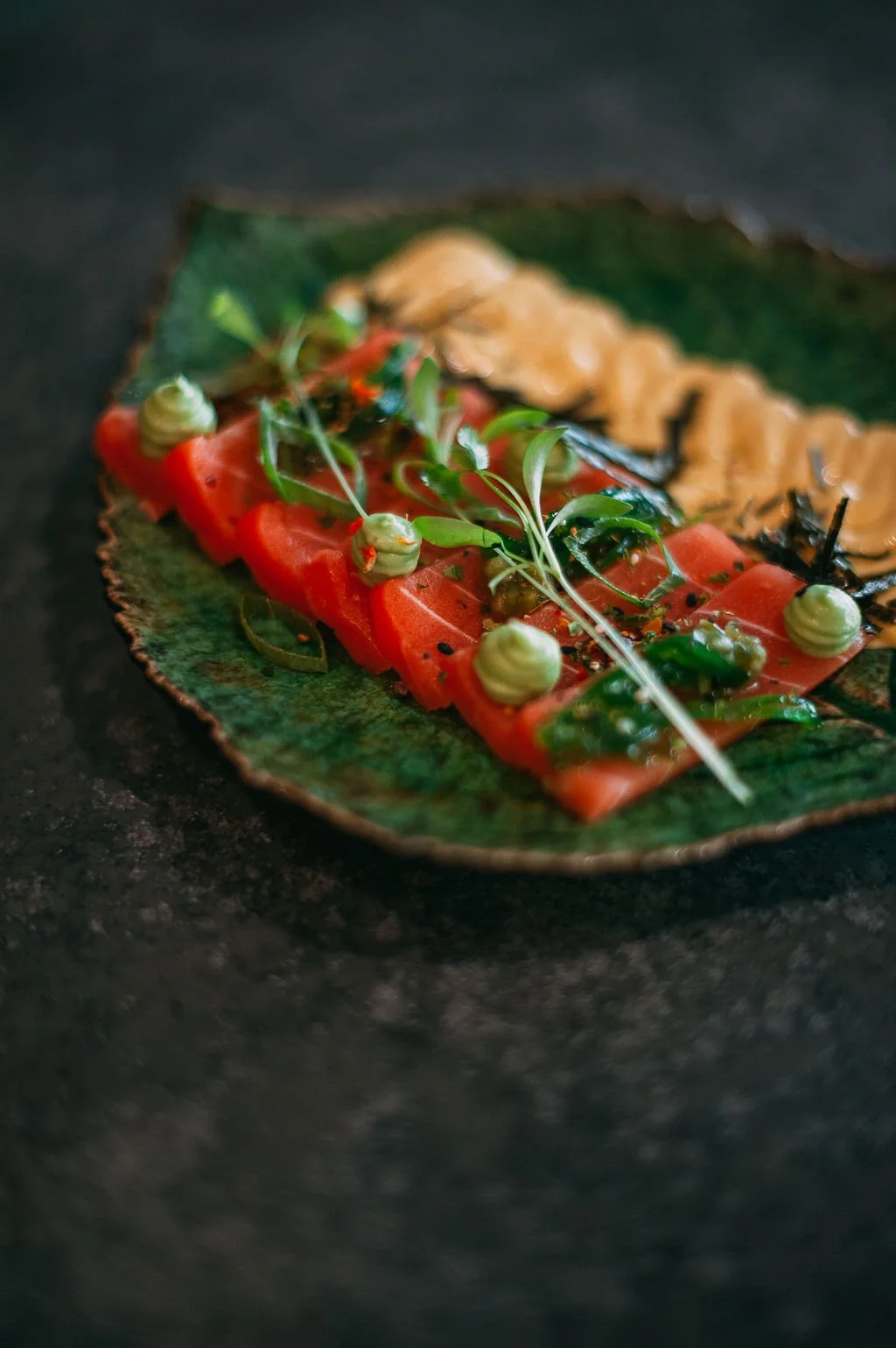 Close-up of a rectangular piece of smoked salmon topped with microgreens, dollops of green mousse, black sesame seeds, and thin slices of avocado, served on a green leaf-shaped ceramic plate.