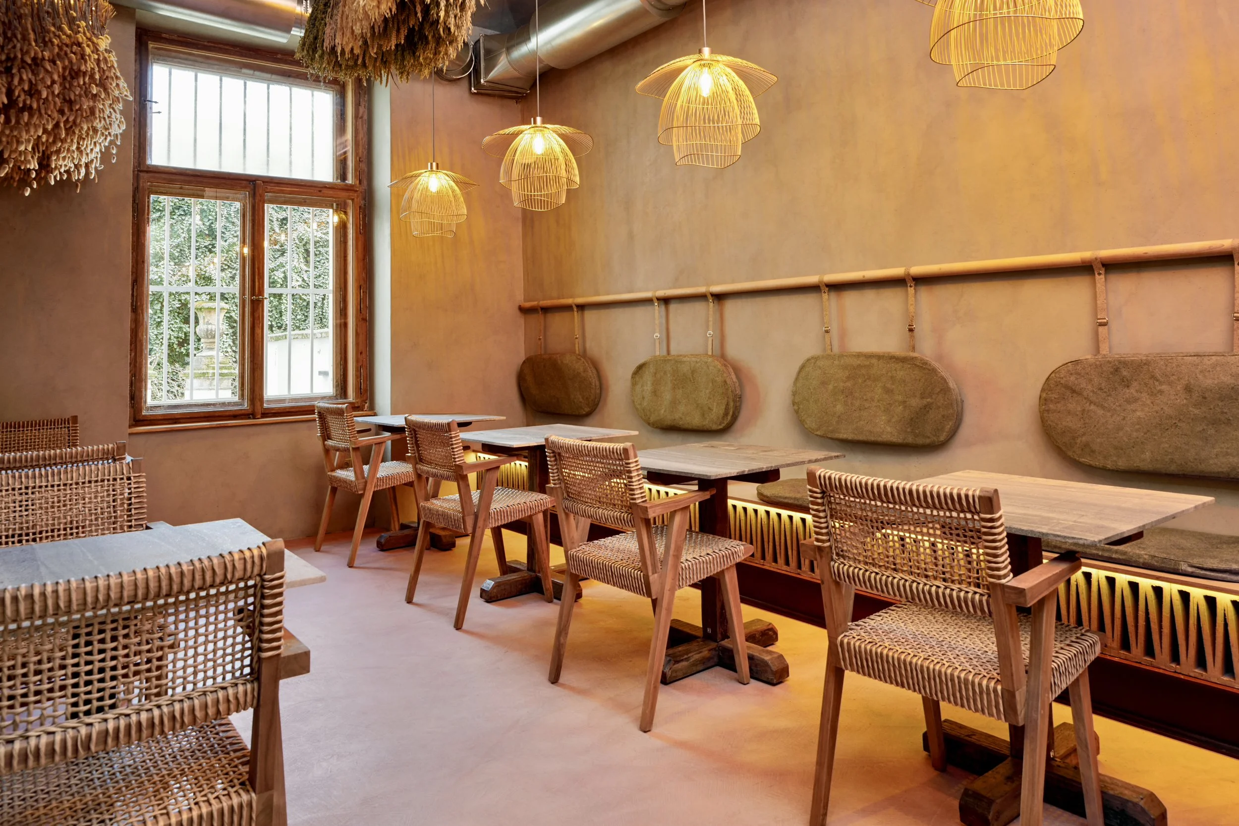 Interior of a cozy cafe with wooden tables and chairs, warm lighting fixtures, a large window, and decorative hanging elements on the ceiling.