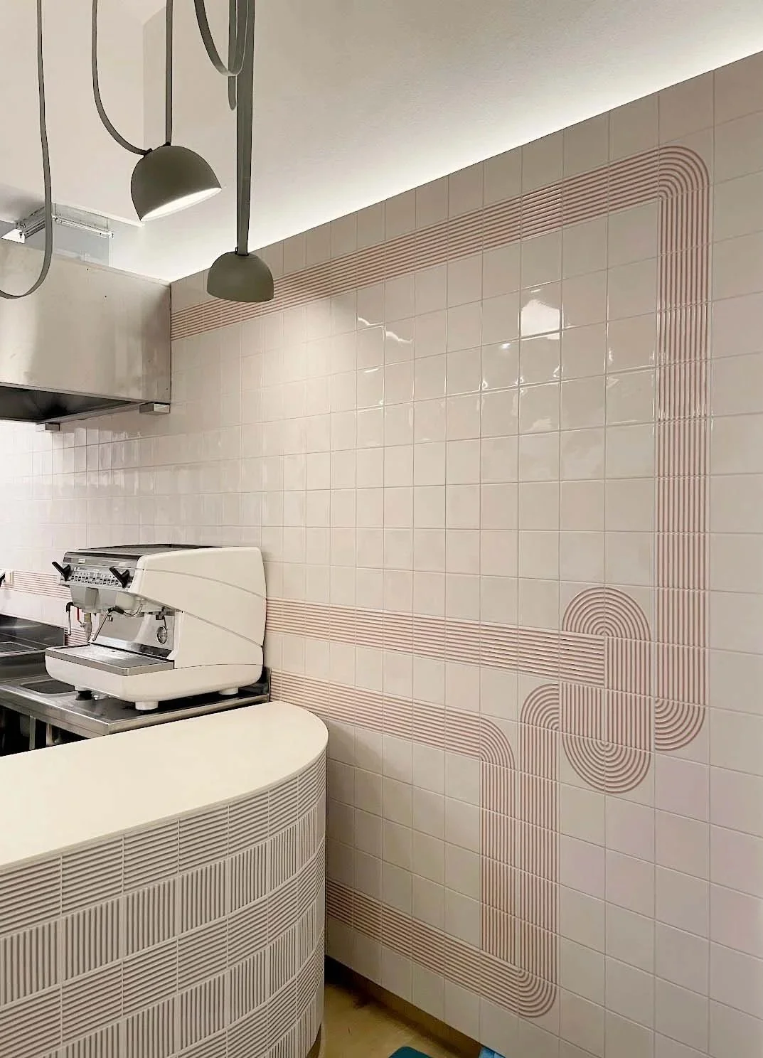 Interior of a retail or cafe space with a tiled wall featuring geometric line patterns in shades of pink and beige, a coffee machine on a curved counter, and industrial-style pendant lights hanging from the ceiling.