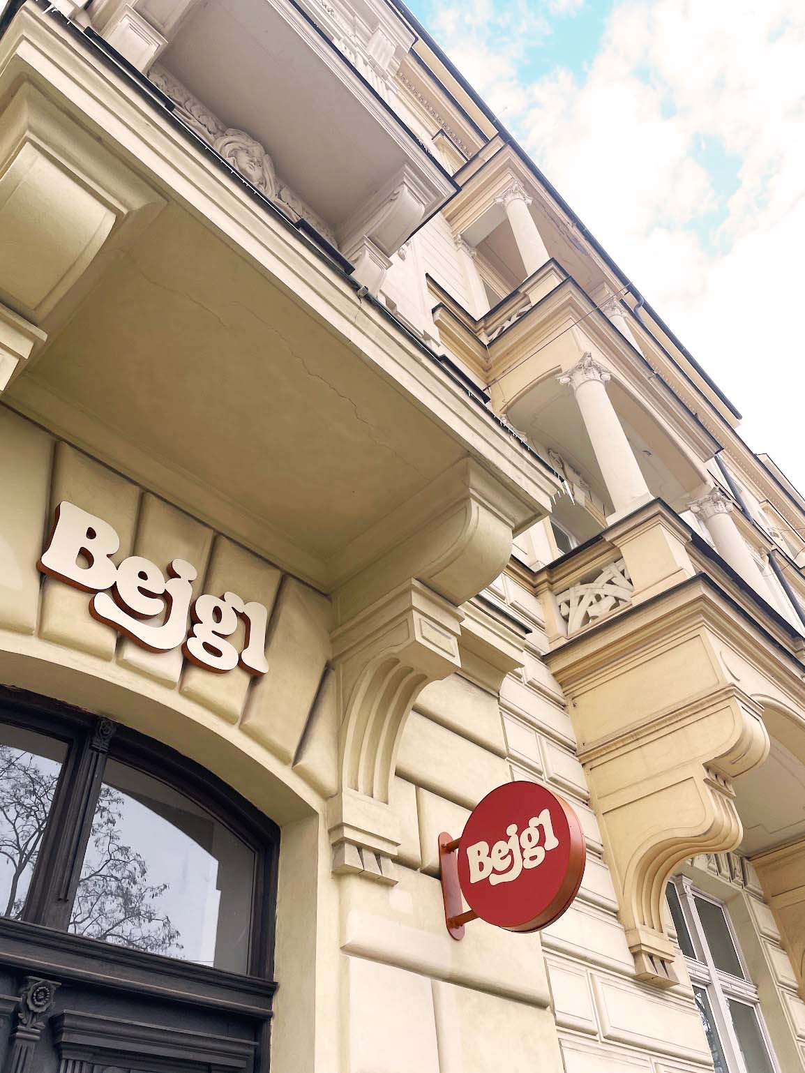 View of a building with ornate architectural details and balconies, featuring a sign with stylized white text on a red circular background that reads 'Begl'.
