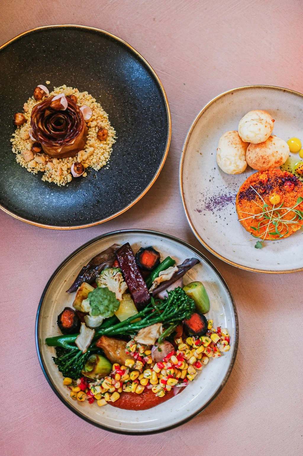 Three plated dishes of gourmet food with vegetables, potato balls, and a dessert featuring a rose-shaped chocolate with crumbles and nuts.