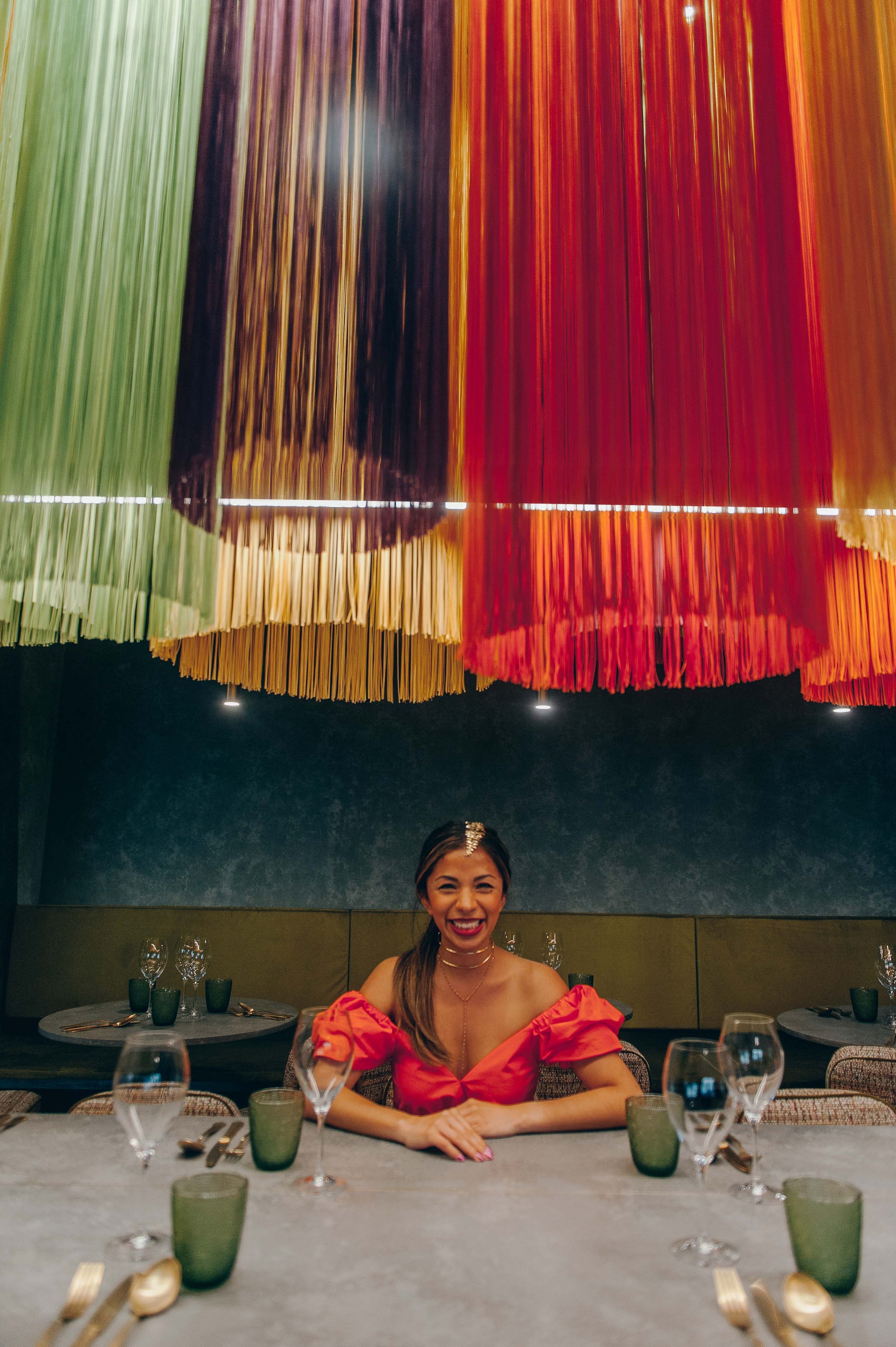 A woman in an orange dress with puffed sleeves and gold jewelry, smiling and sitting at a dinner table set with glasses and utensils, in a restaurant with colorful hanging decorations on the ceiling.