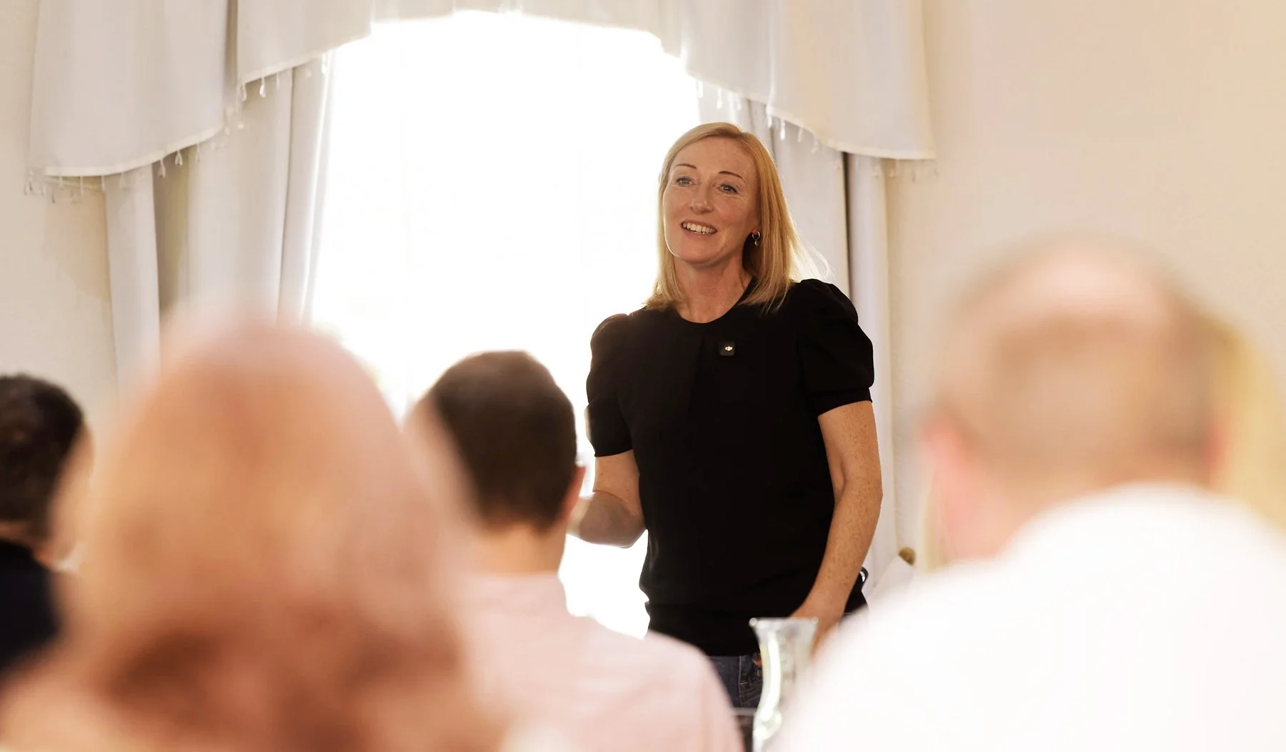 Smiling woman in black speaking to a seated audience in a bright room